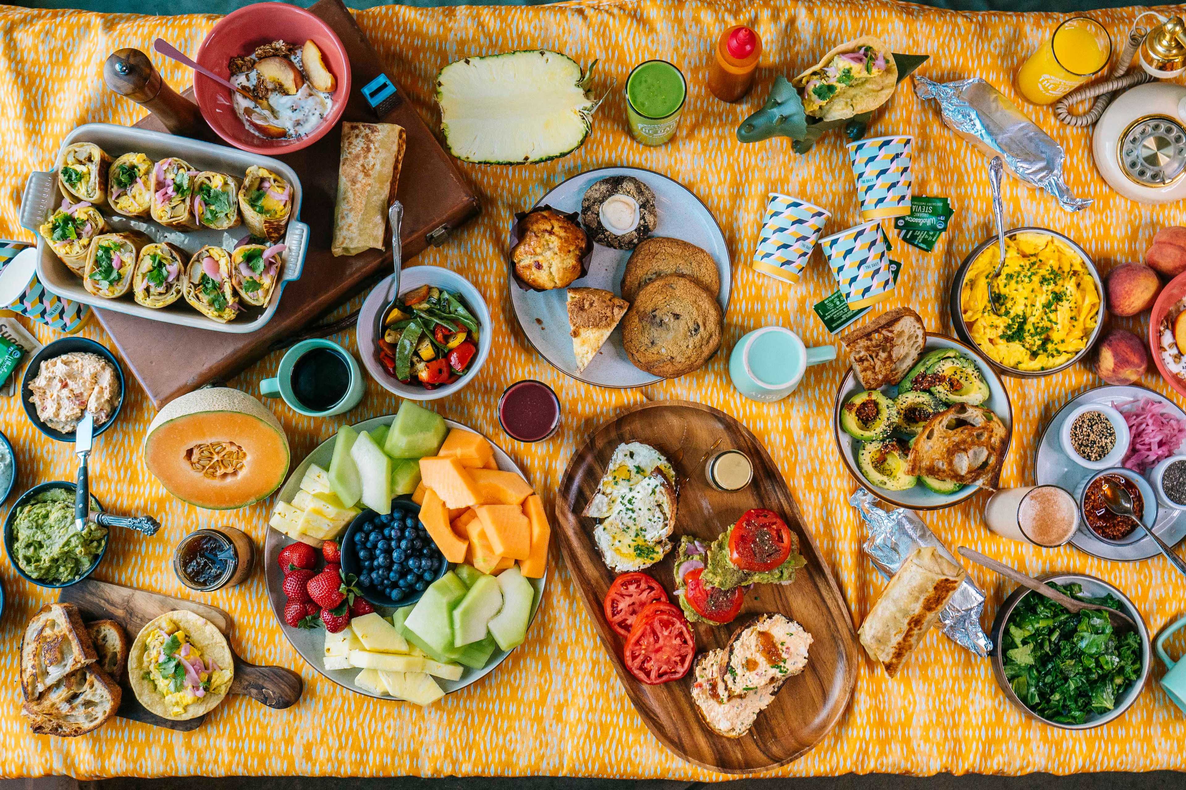 The image displays a variety of colorful food items arranged on a patterned yellow tablecloth, including fruits, sandwiches, salads, and baked goods.