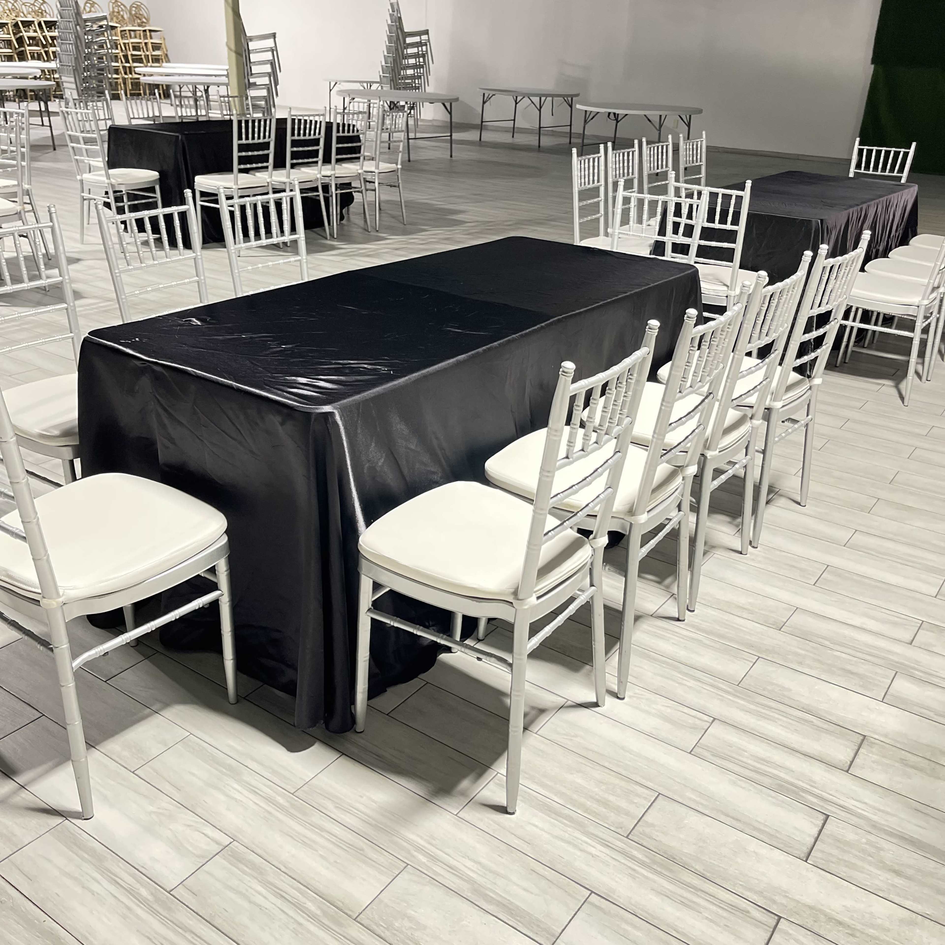 A large room features several tables covered in black and white linens, surrounded by white chairs arranged neatly on a tiled floor.