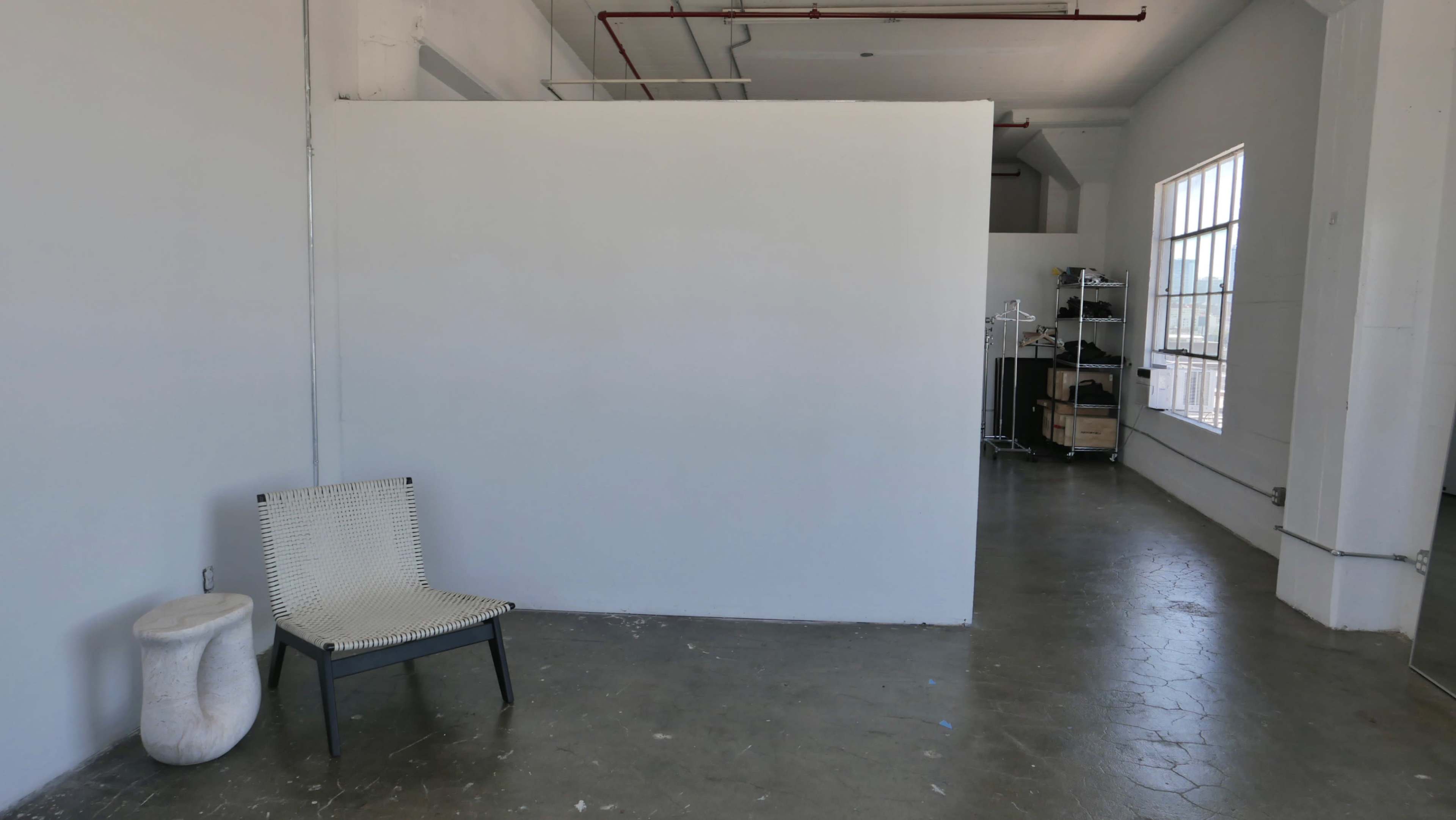 The image depicts a spacious, minimalist interior with a plain white wall, a simple chair on the left, and shelves in the background.