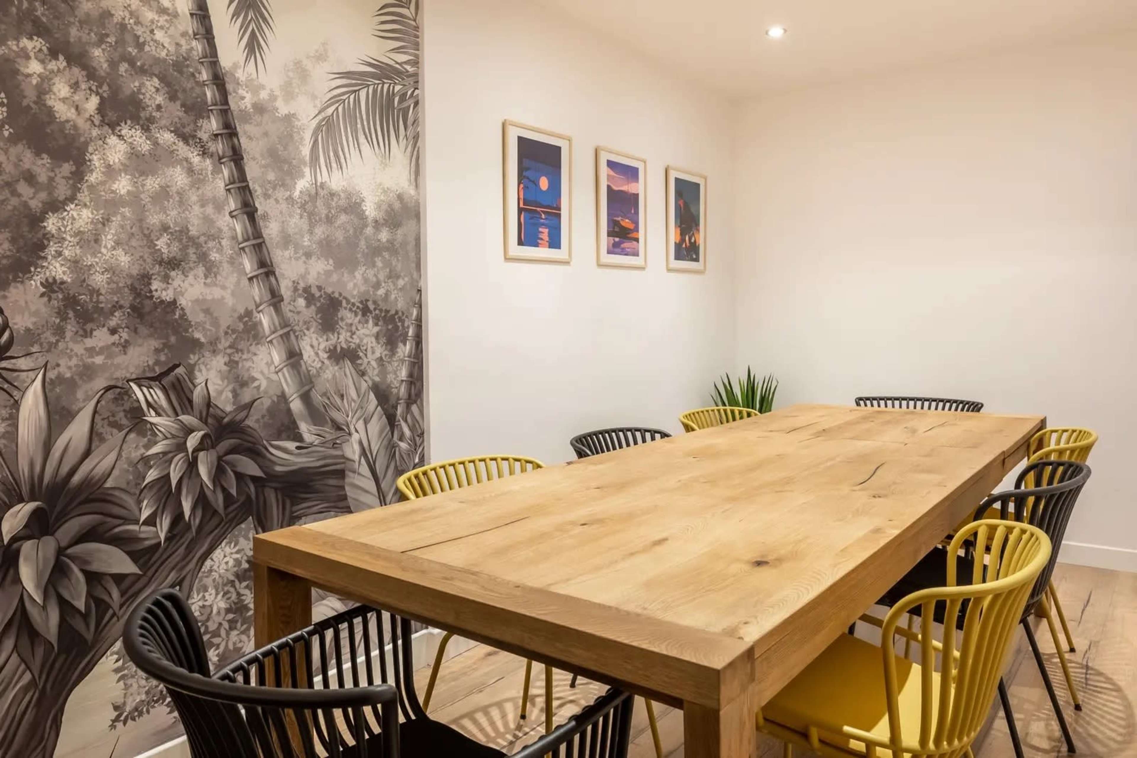 The image shows a modern dining room with a large wooden table surrounded by black and yellow chairs, against a backdrop of a nature-themed mural and framed artwork on the walls.