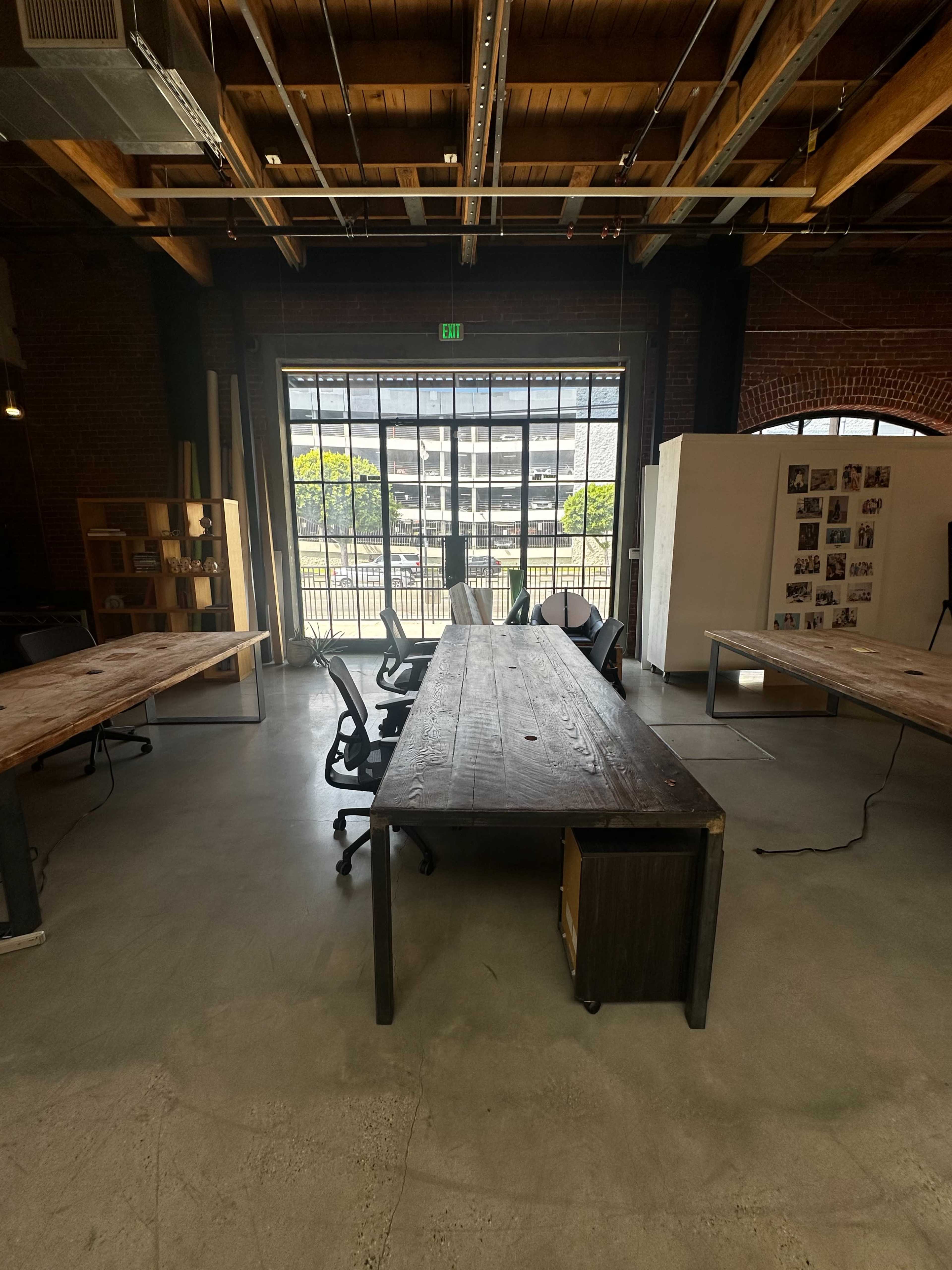A large wooden conference table is centered in an open workspace with several office chairs and large windows offering a view of the outside.