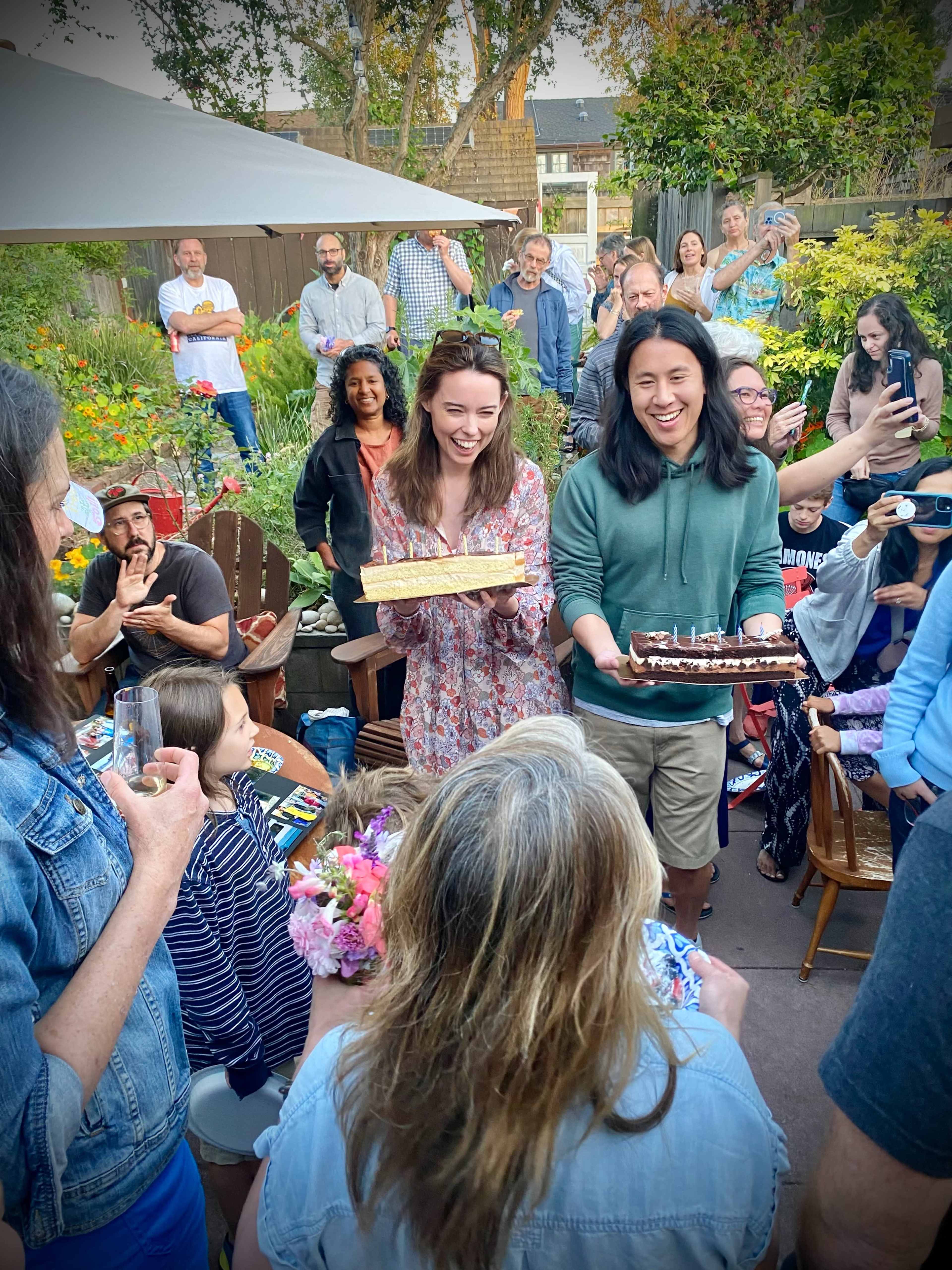 A group of people gathers in a backyard to celebrate with two individuals carrying cakes while others take photos and share the moment.