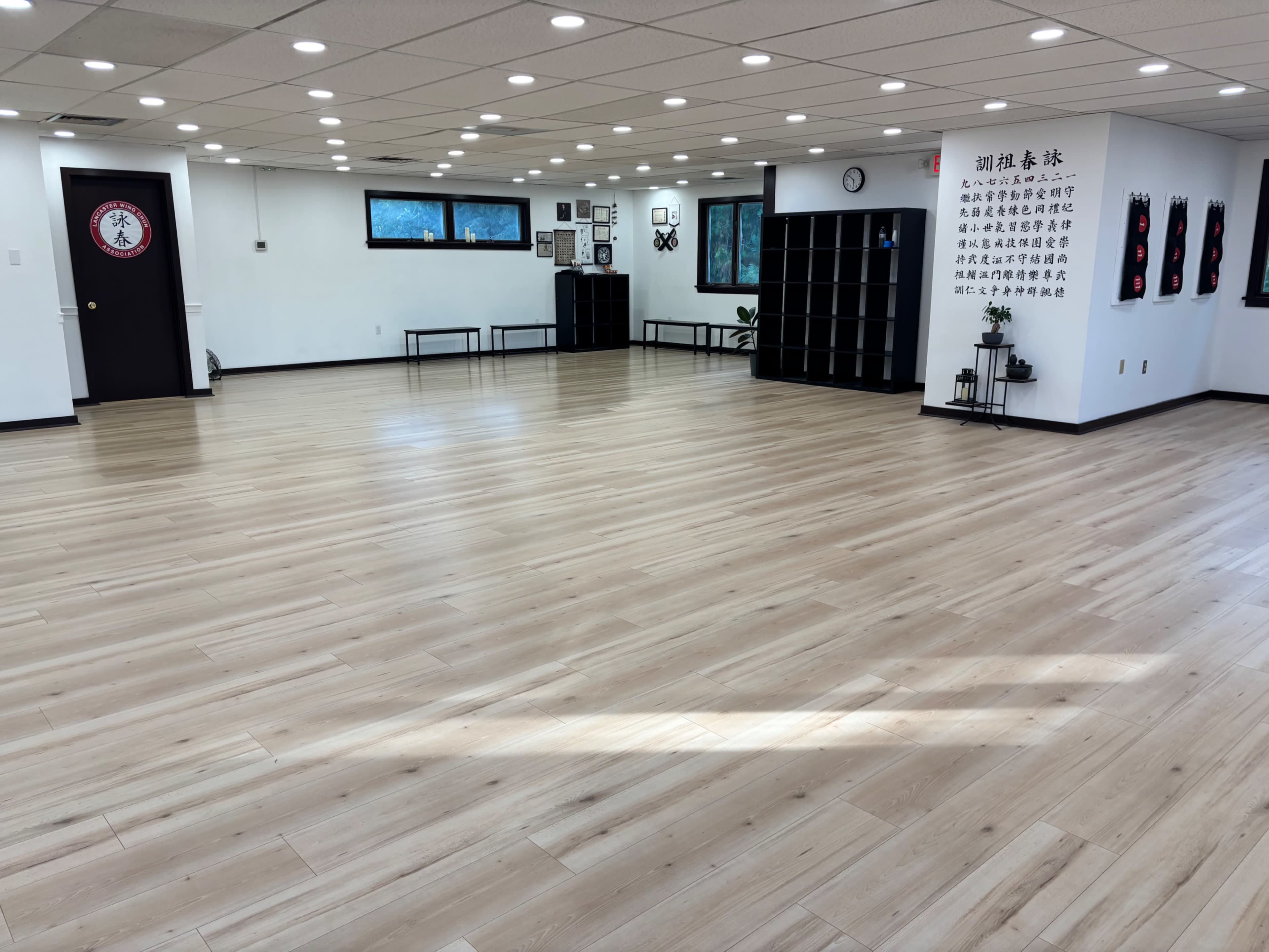 The image shows an empty, well-lit studio space with wooden flooring and minimalistic decor, including shelves and wall art.
