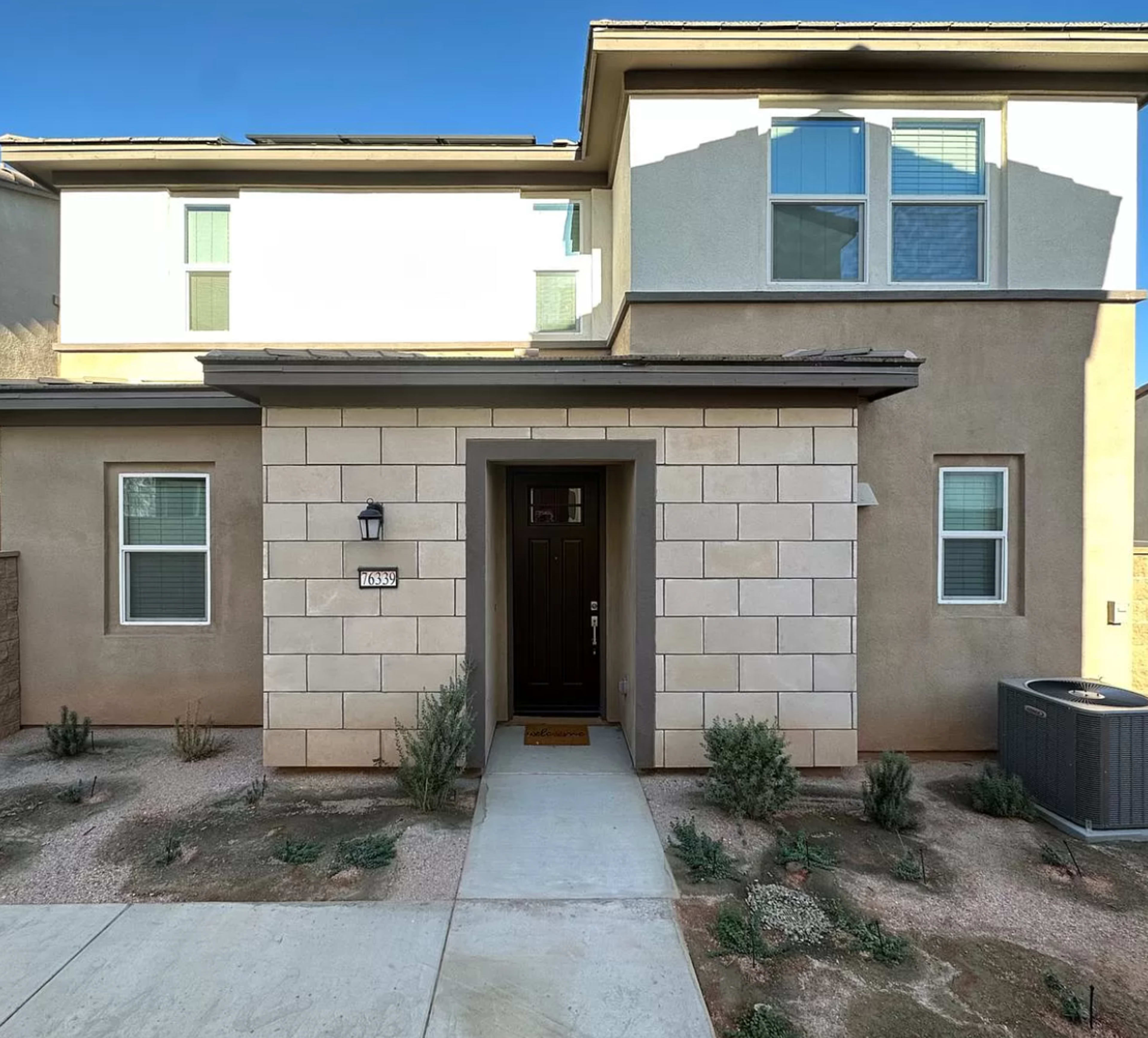 The image shows the front entrance of a modern two-story home with a light-colored facade, a dark door, and a small pathway leading to it.
