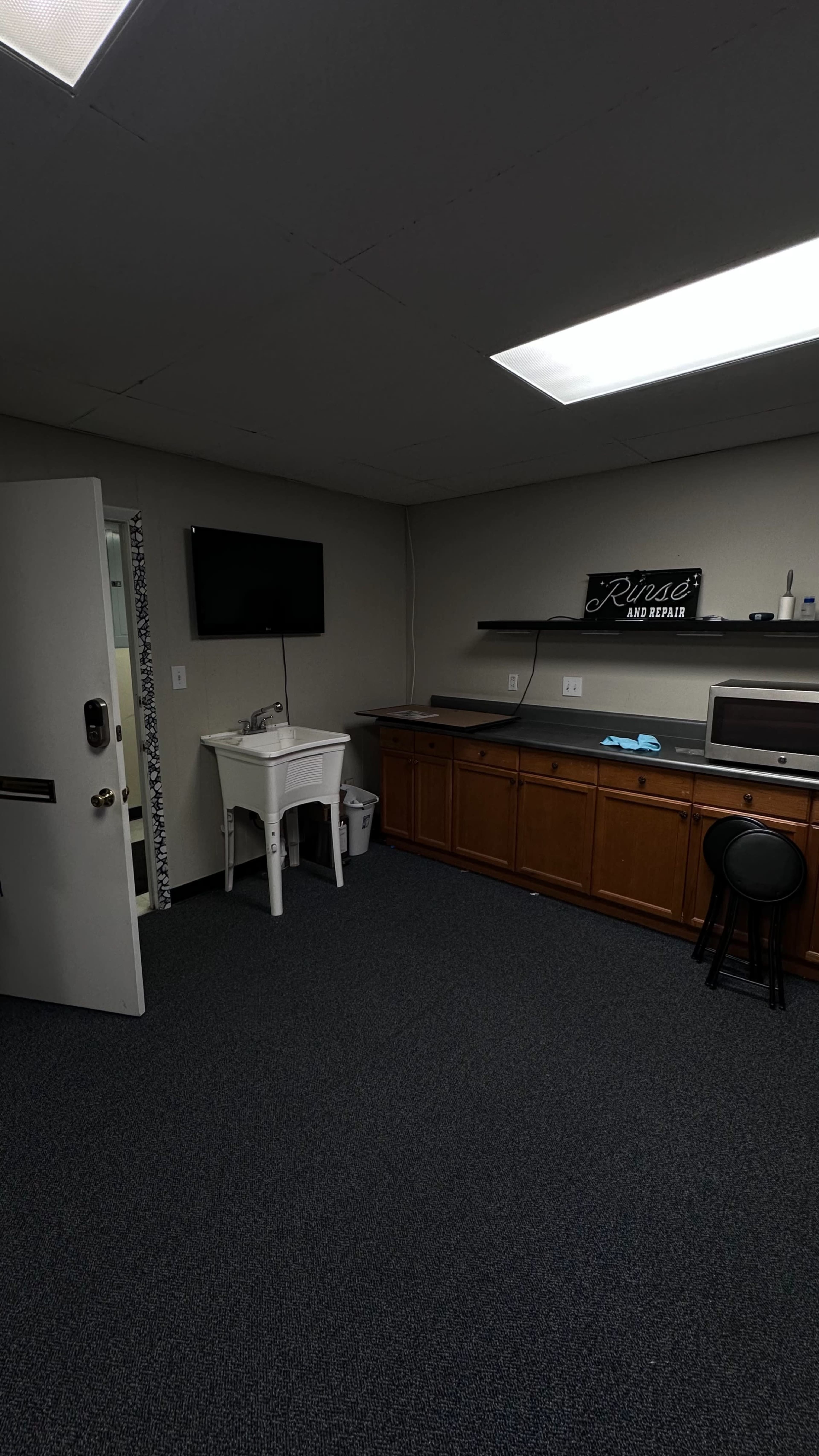 The image shows a small office or workspace featuring a wooden counter, a sink, a stool, and a wall-mounted television.