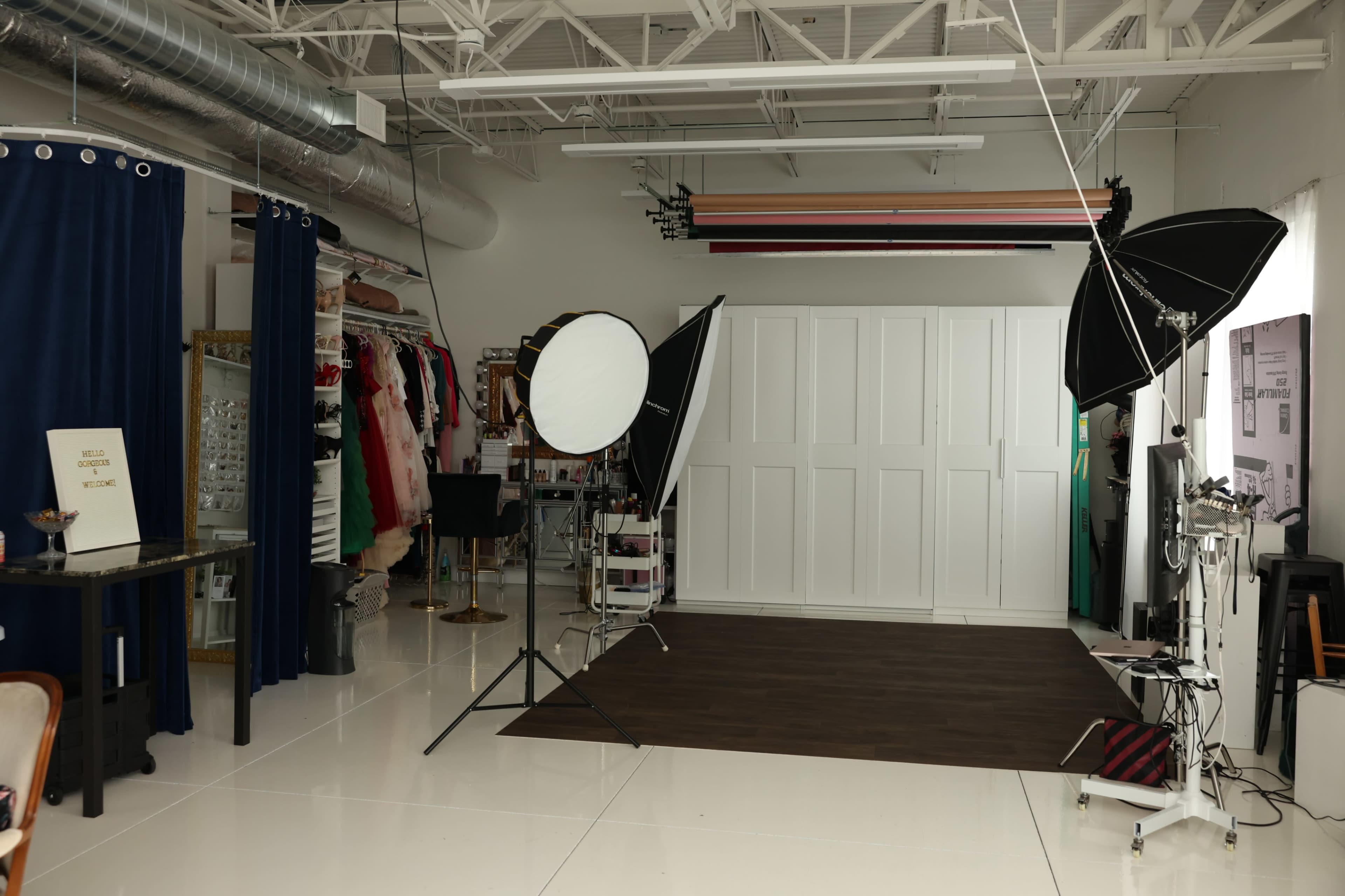 The image shows a photography studio with a white backdrop, lighting equipment, and various clothing items hanging on racks.