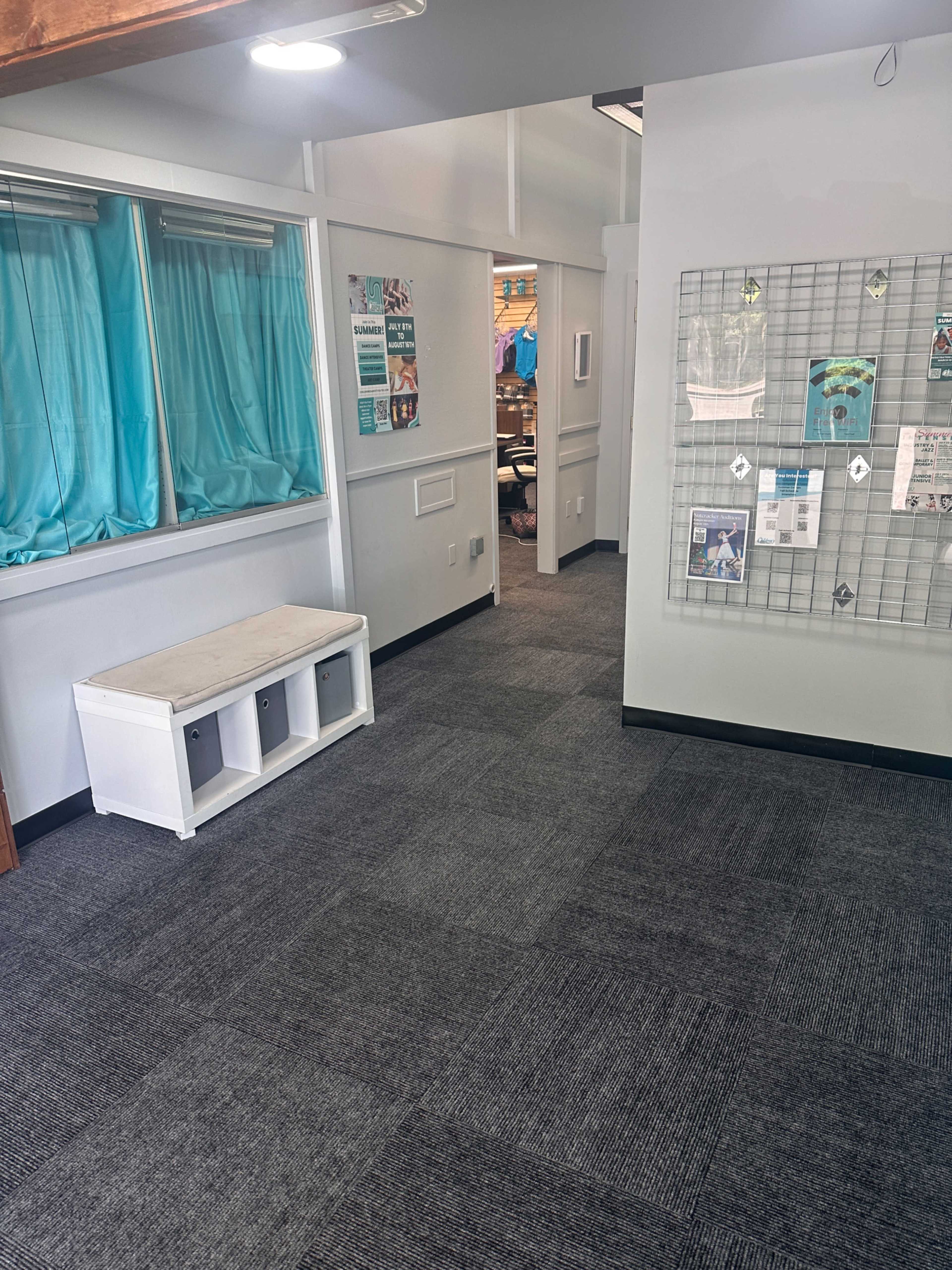 The image shows a well-lit interior space with gray carpet, a bench with storage cubbies, and a hallway leading to another room, adorned with wall-mounted informational displays.