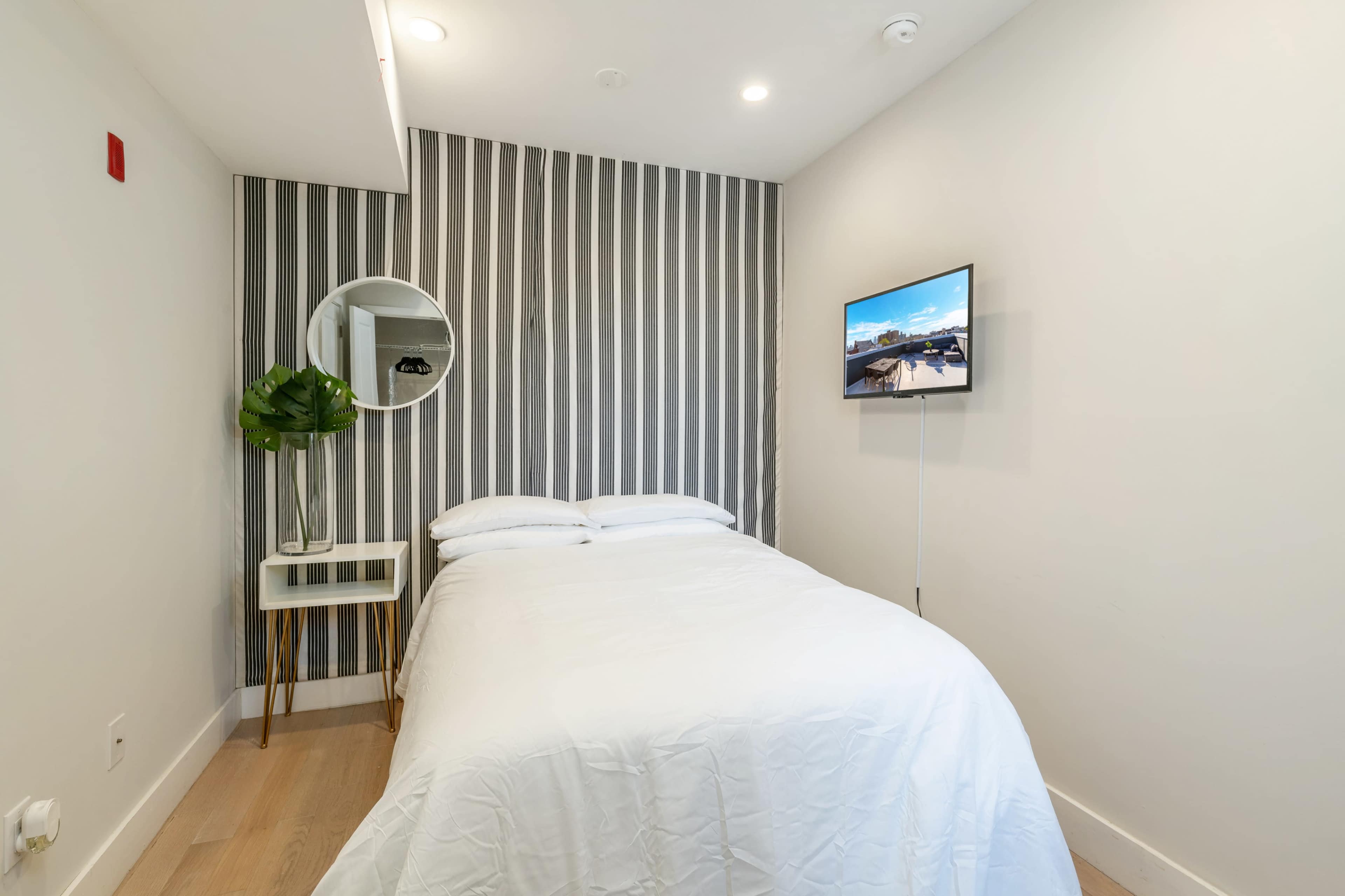 A small, minimalist bedroom featuring a bed, a round mirror, a potted plant, and a mounted television on the wall.