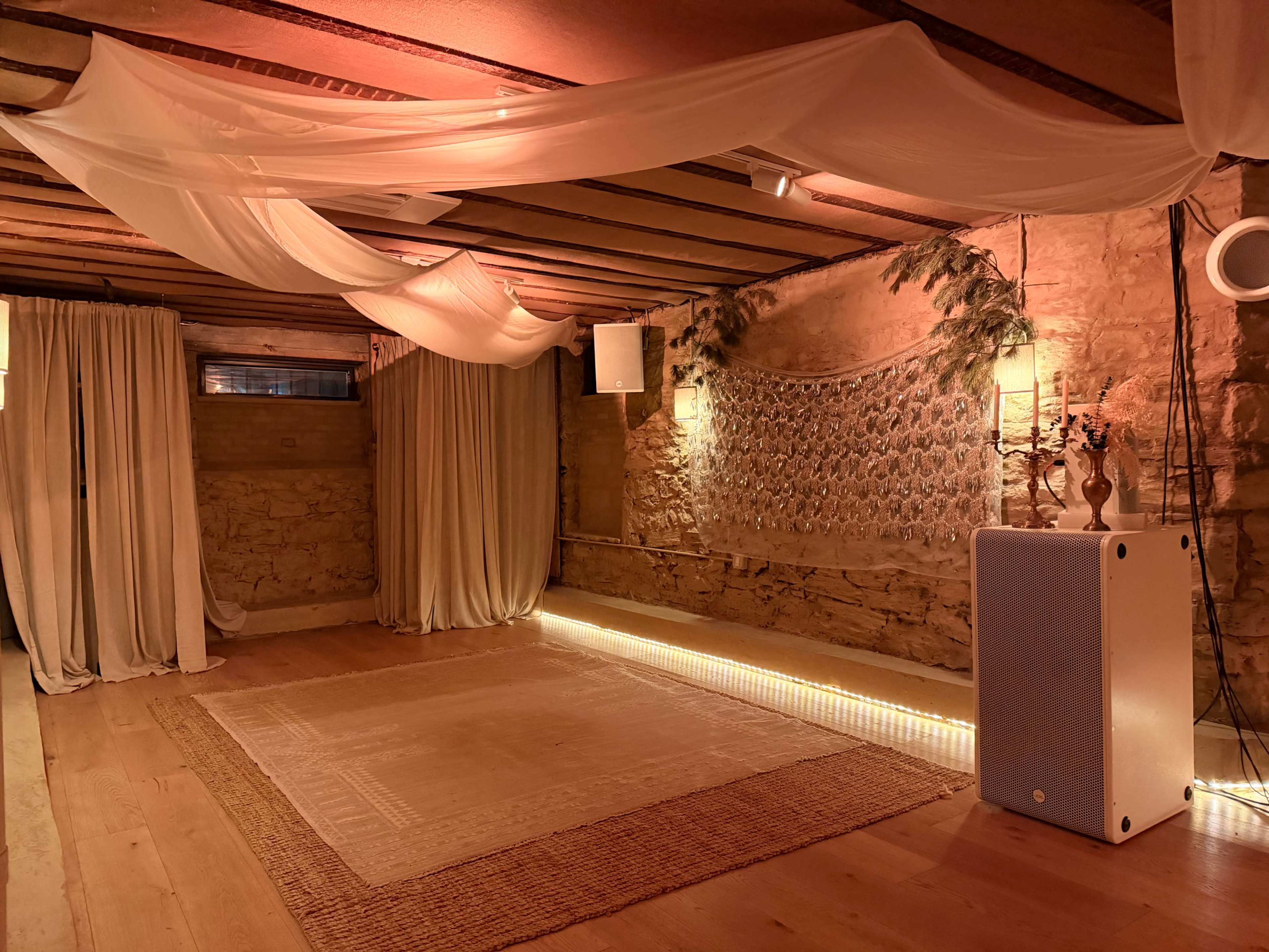 The dimly lit room features wooden beams, flowing fabric drapes, and a textured stone wall with soft, ambient lighting along the floor.