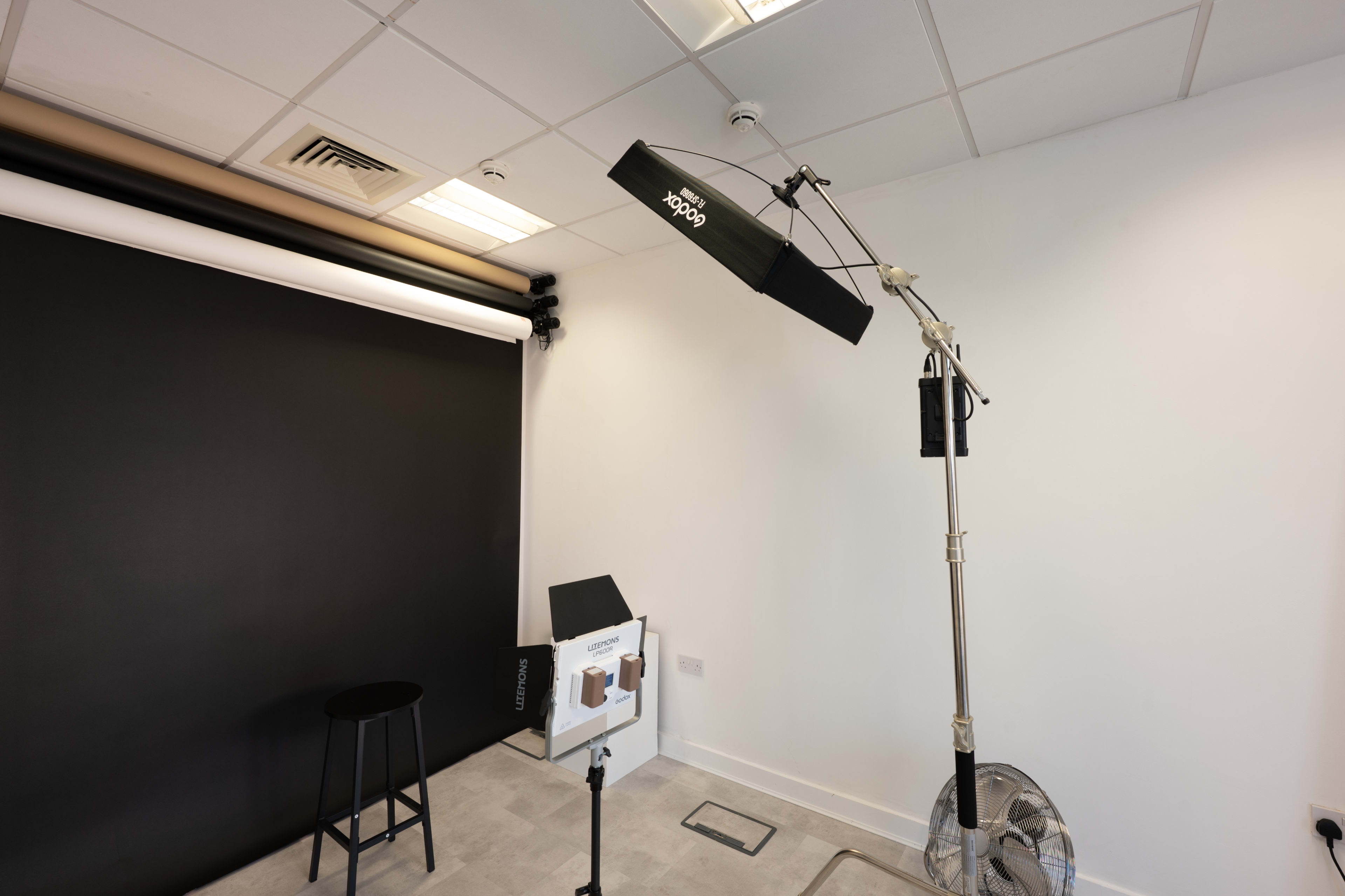 The image shows a photography studio with a black backdrop, lighting equipment mounted on a stand, a stool, and a fan on the floor.