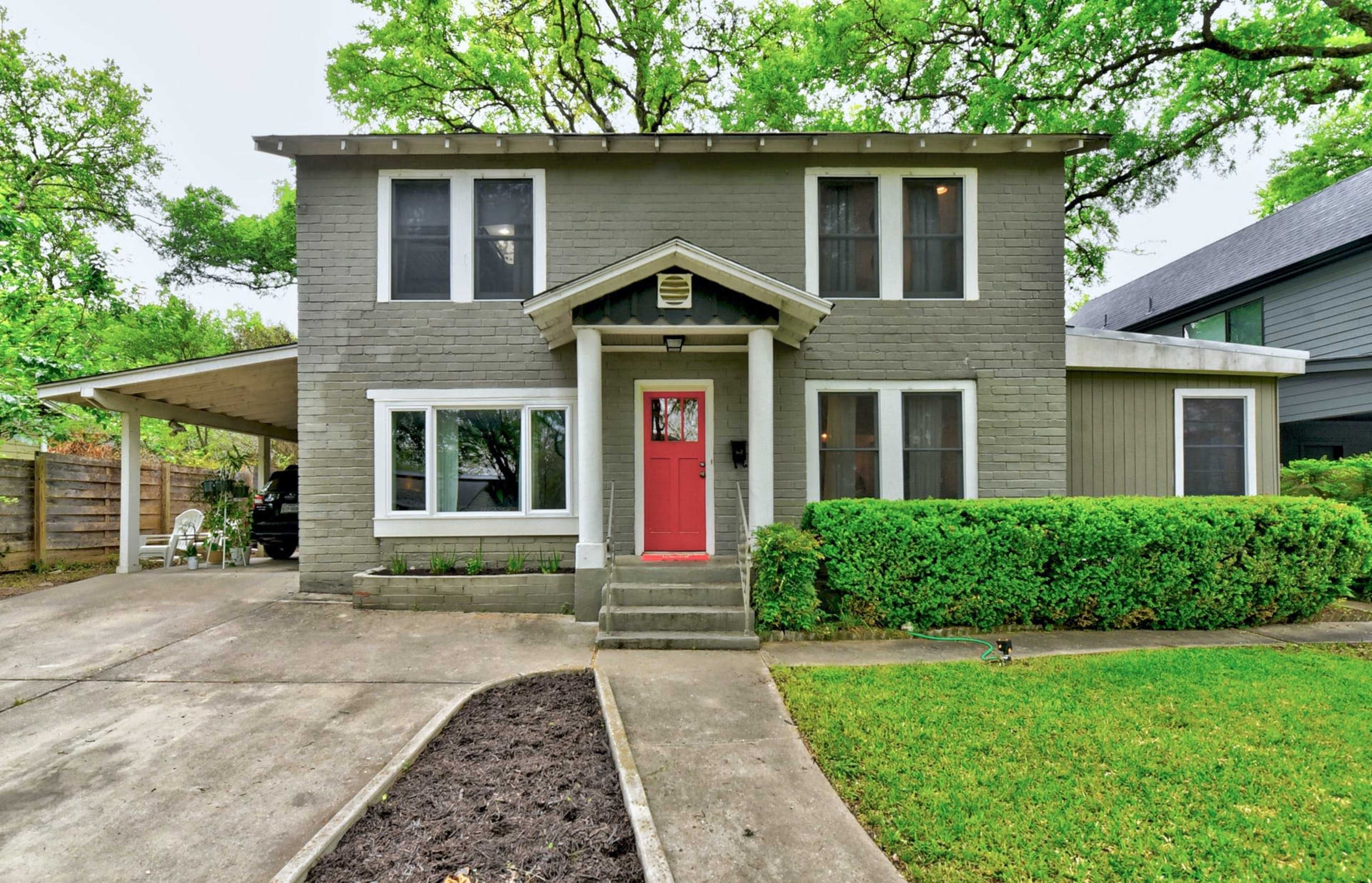 A two-story gray house with a red front door is surrounded by green foliage and has a carport on the left side.