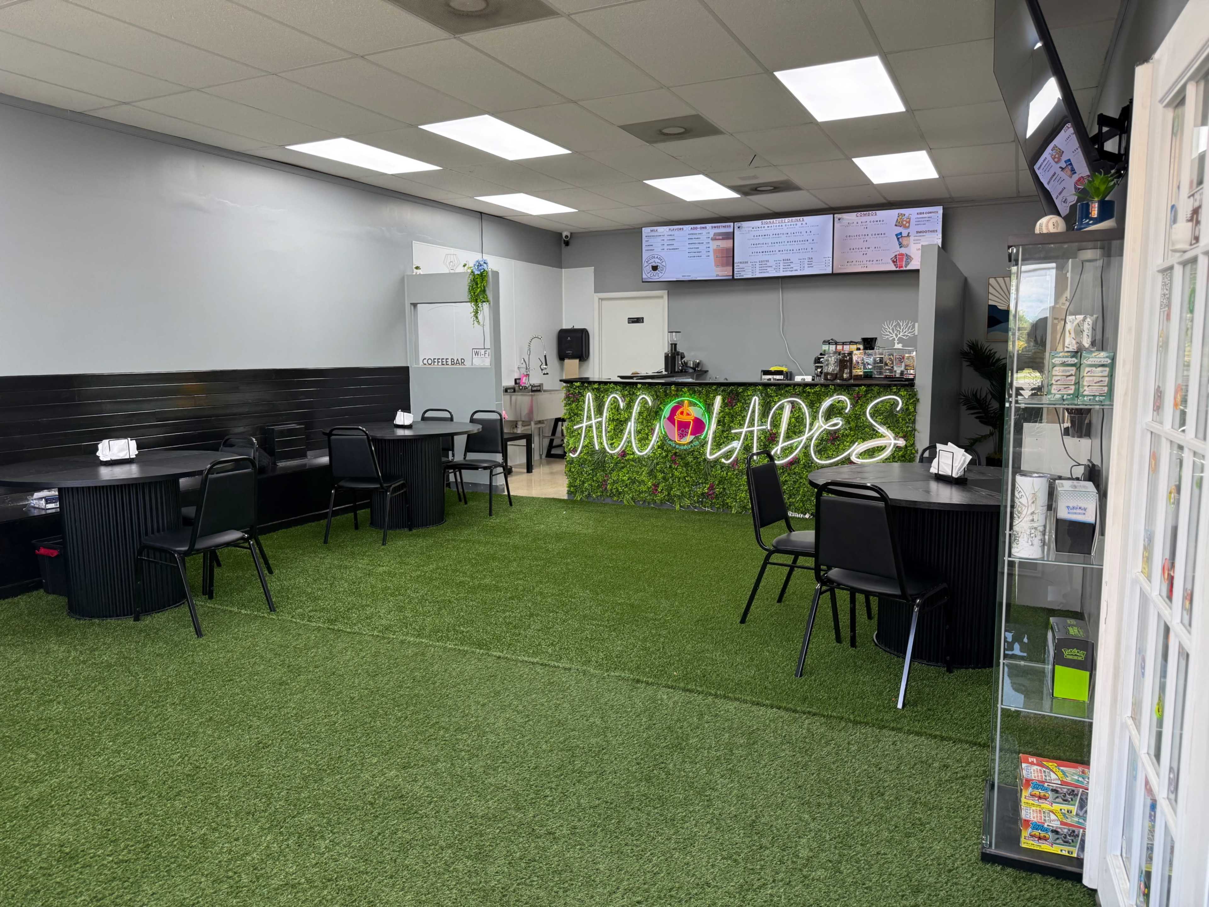 The image shows a clean and modern café interior with artificial grass flooring, several tables with chairs, and a counter displaying a menu.