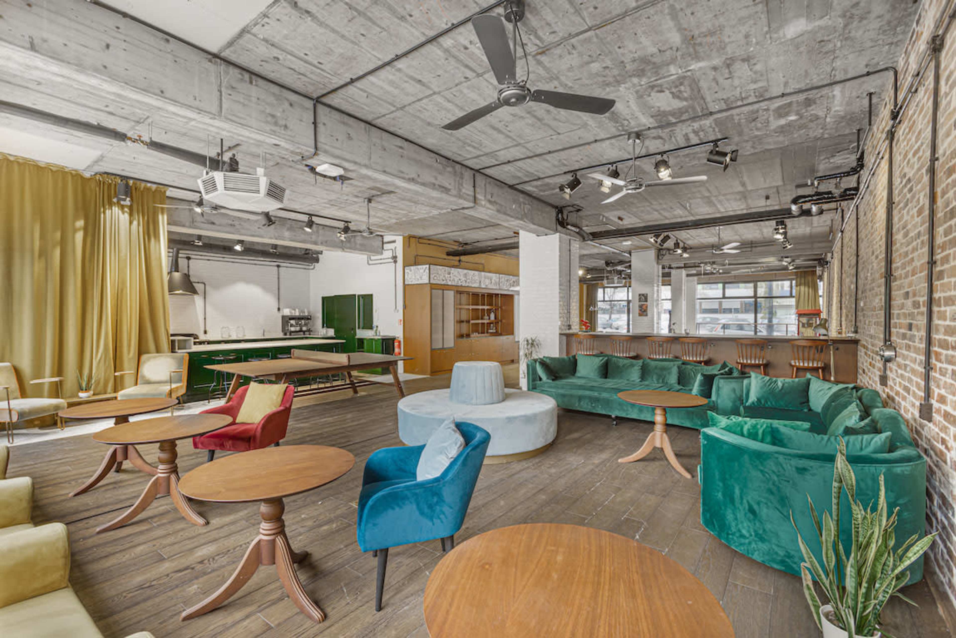 The image shows a spacious modern interior with circular seating arrangements, wooden tables, and a kitchen area in the background.