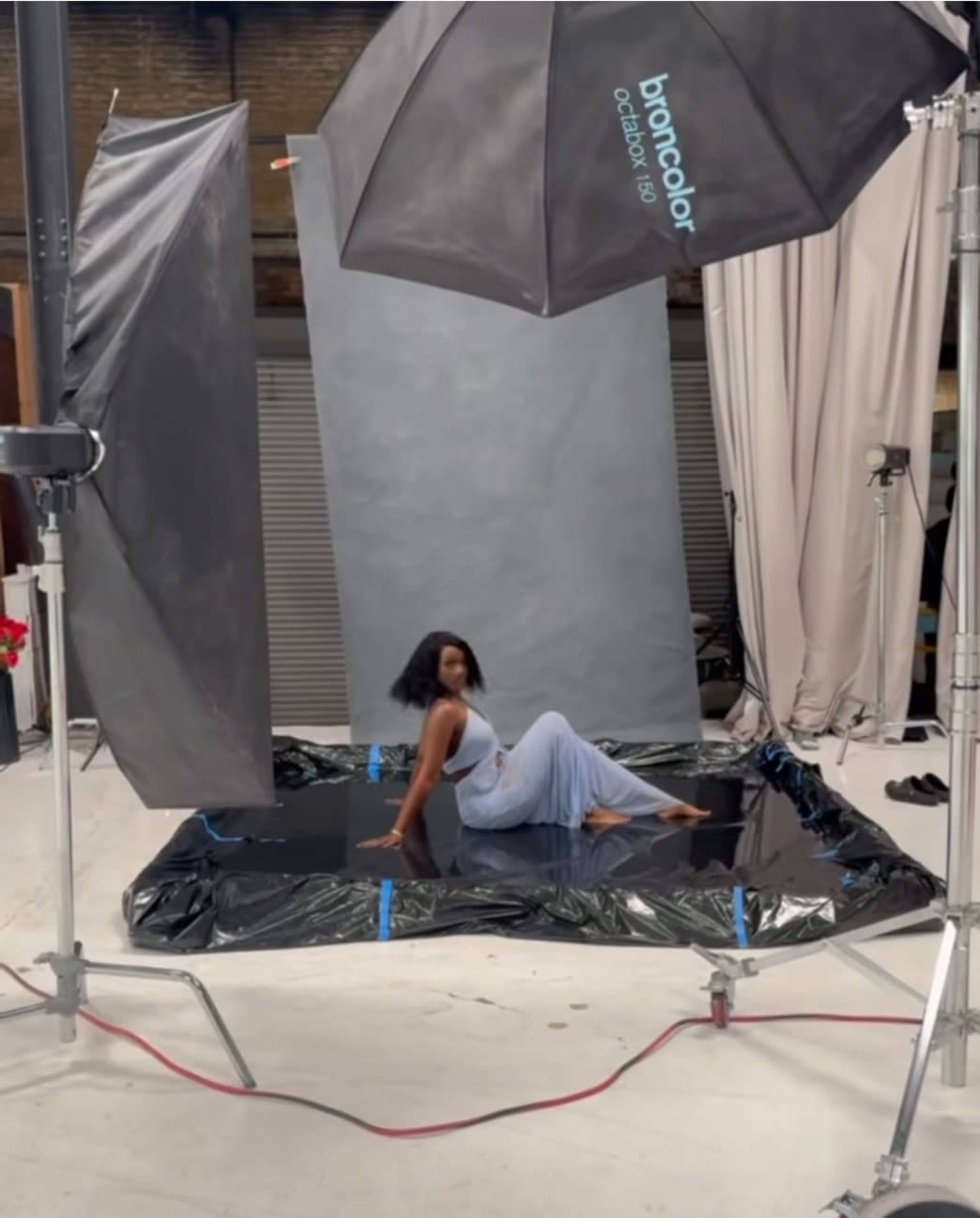 A model poses on a reflective surface in a photography studio, surrounded by large softbox lights and backdrop panels.
