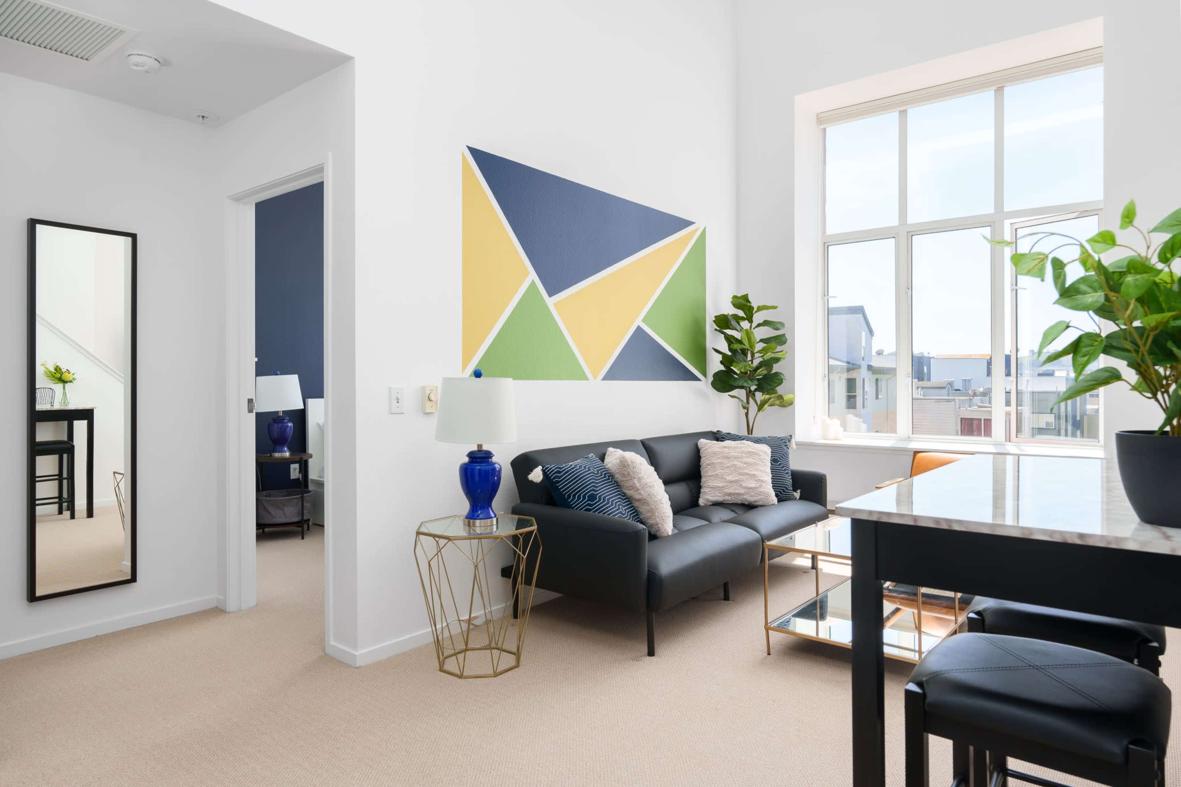 A modern living space features a black sofa, a small side table with a lamp, and a colorful abstract painting on the wall beside a large window with potted plants.