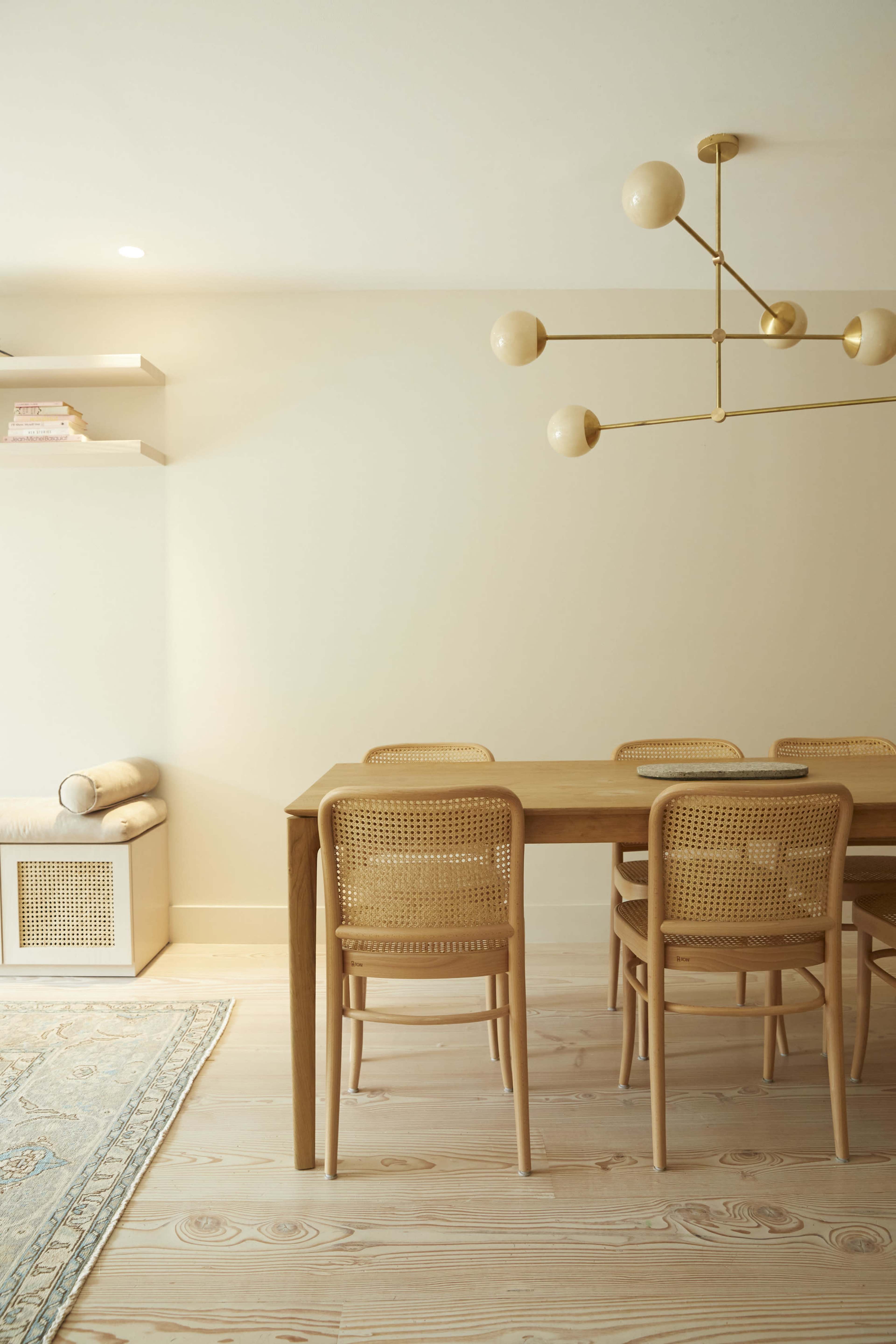 A wooden dining table with six cane chairs is positioned in a softly lit room, accompanied by a light fixture above and shelves on the wall.