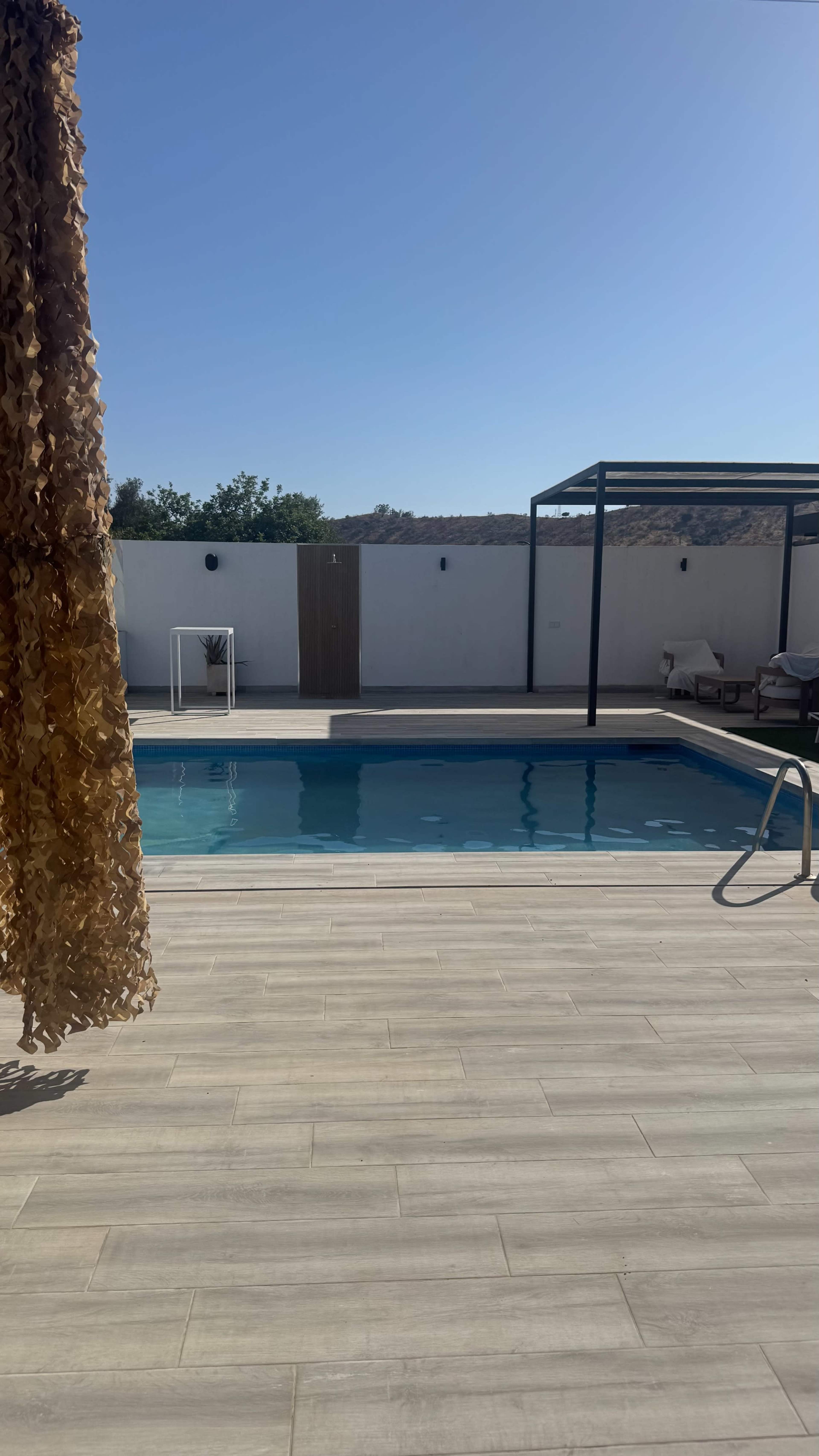 A clear swimming pool is located within a private outdoor area featuring a shaded lounging space and a wooden deck.