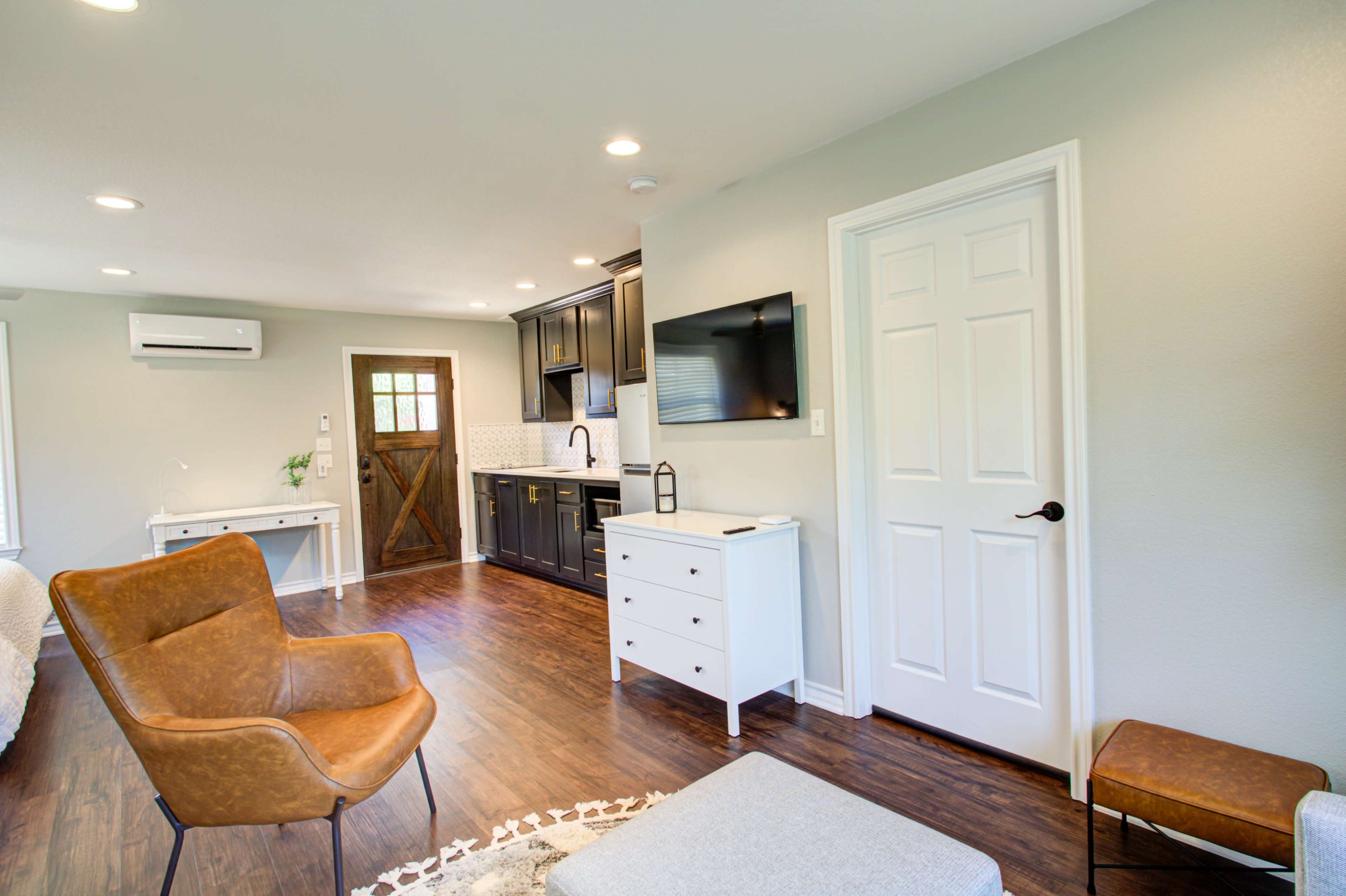 A modern living space featuring a kitchenette, a seating area with a brown leather chair, and a white cabinet beside a door leading outside.