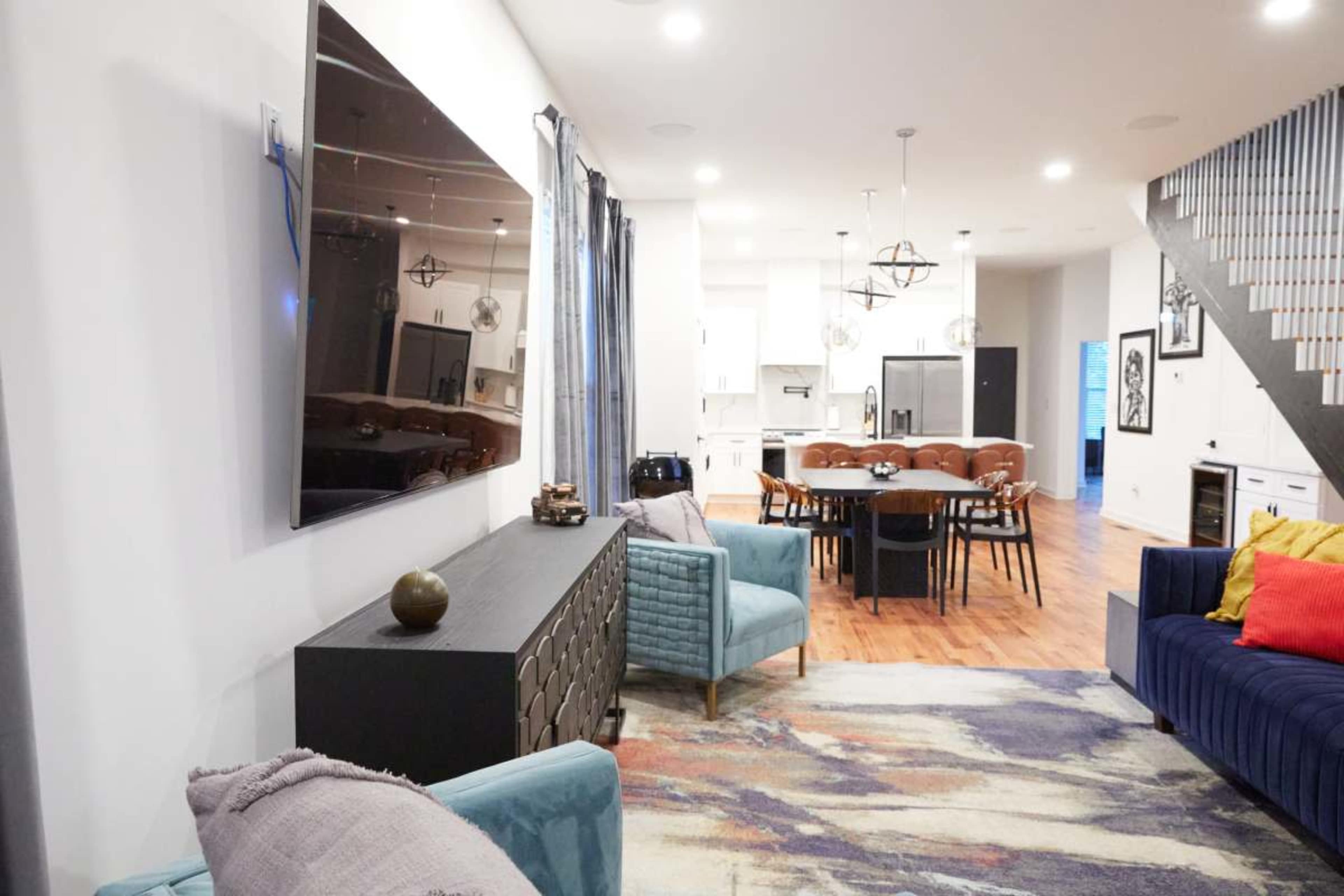 The image shows a modern living space with a television mounted on the wall, a blue sofa, and a dining area featuring a large table and multiple chairs.