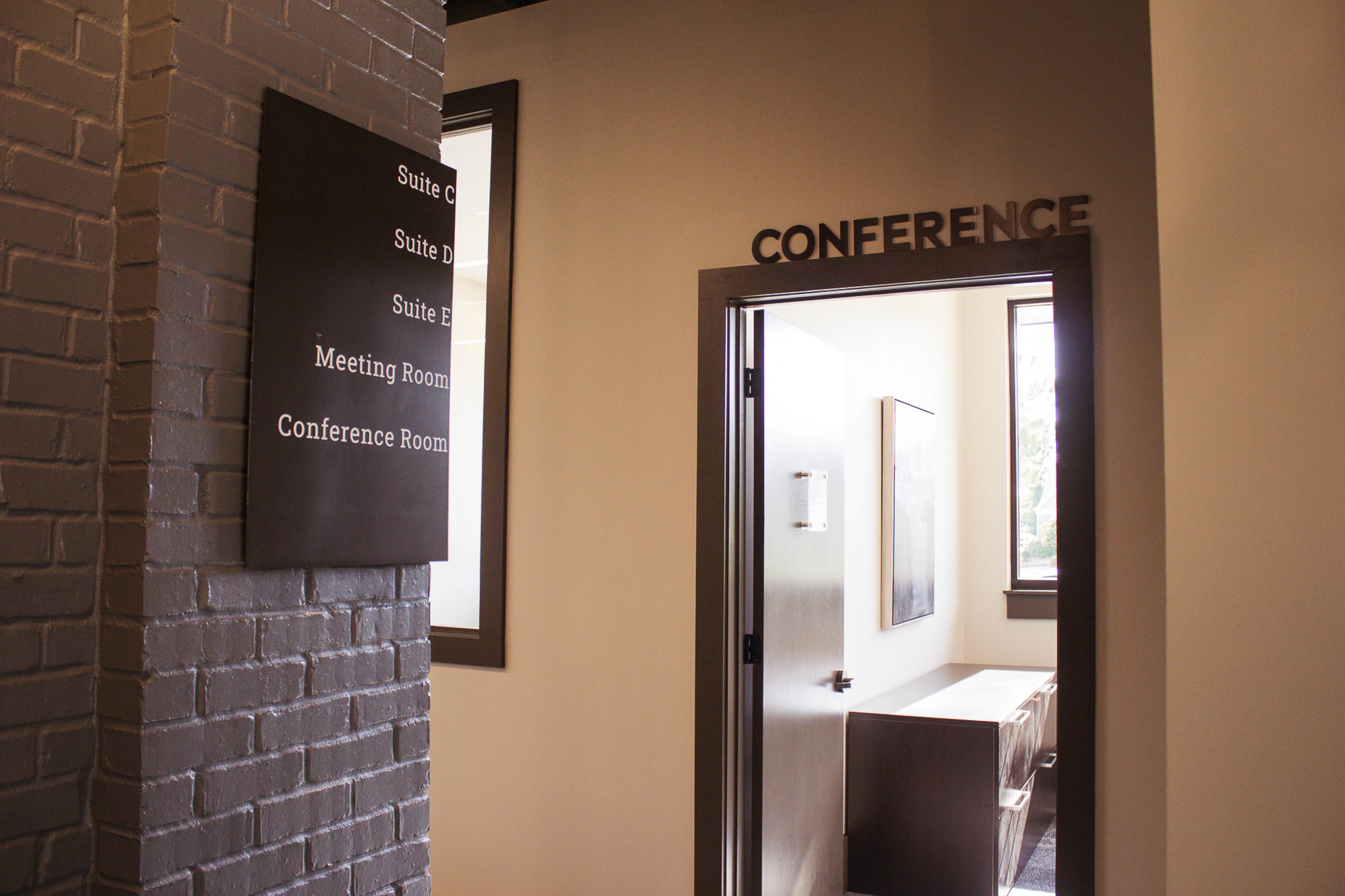 An office corridor with a sign indicating "CONFERENCE" and various suite names on the wall beside an open doorway leading into a conference room.