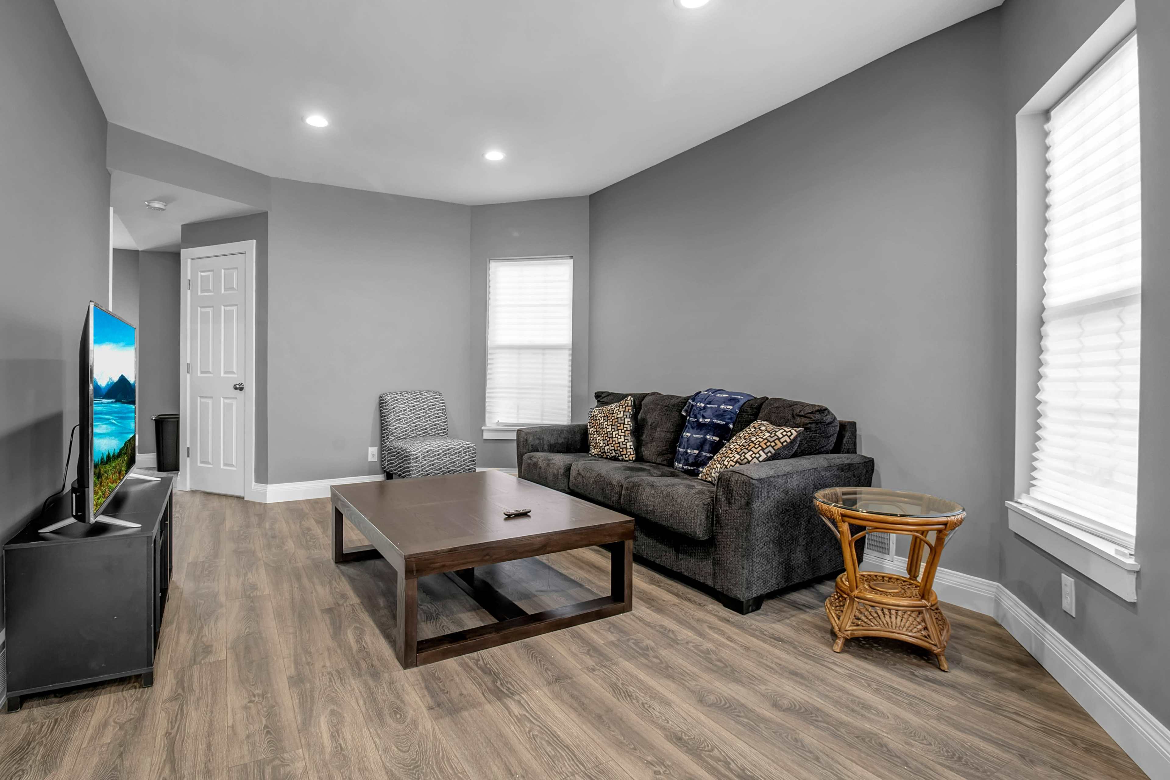 A living room with a dark sofa, a wooden coffee table, a small side table, and a television against a gray wall.
