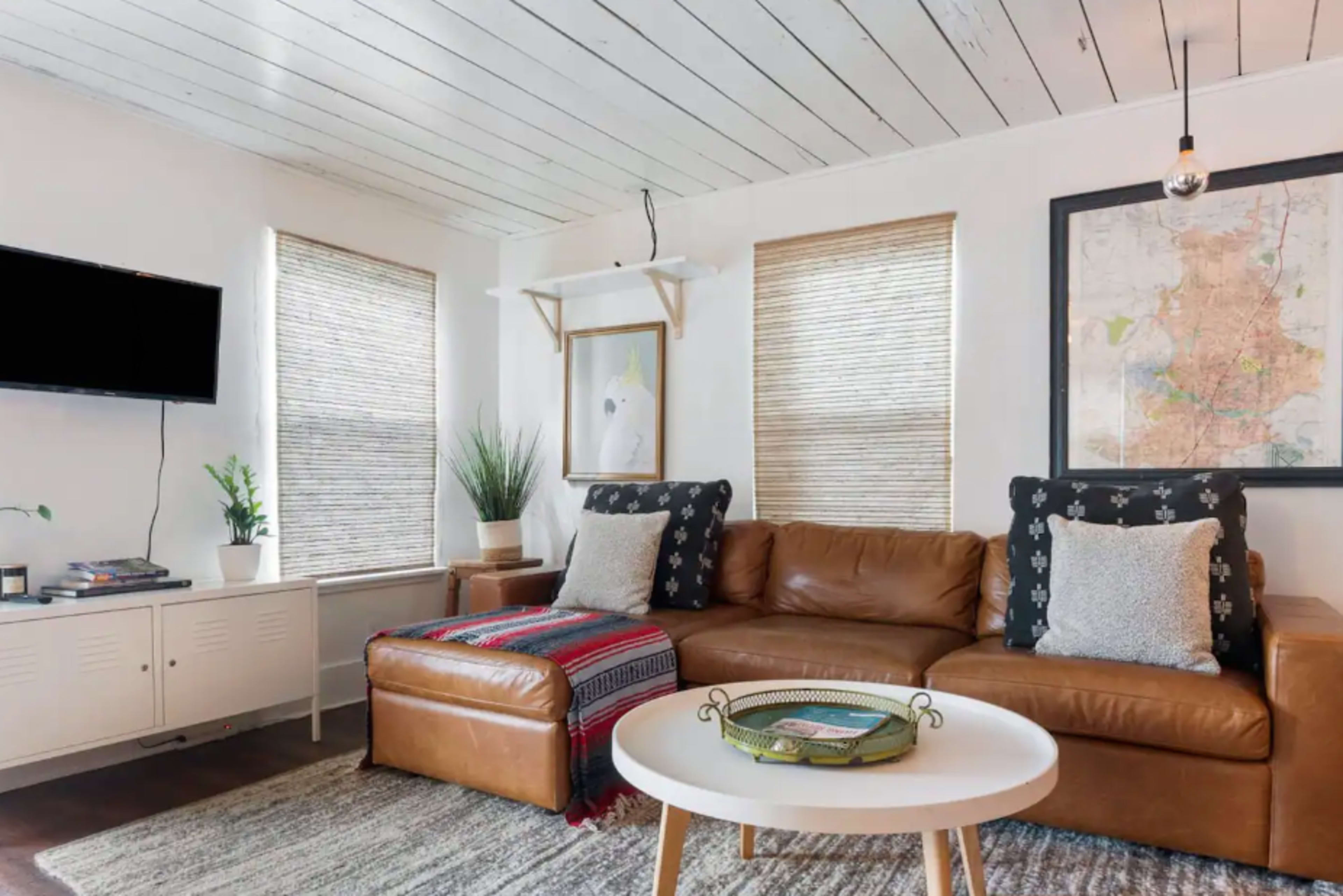 A cozy living room featuring a brown leather sofa, a round table, a wall-mounted television, and plants near large windows with bamboo shades.
