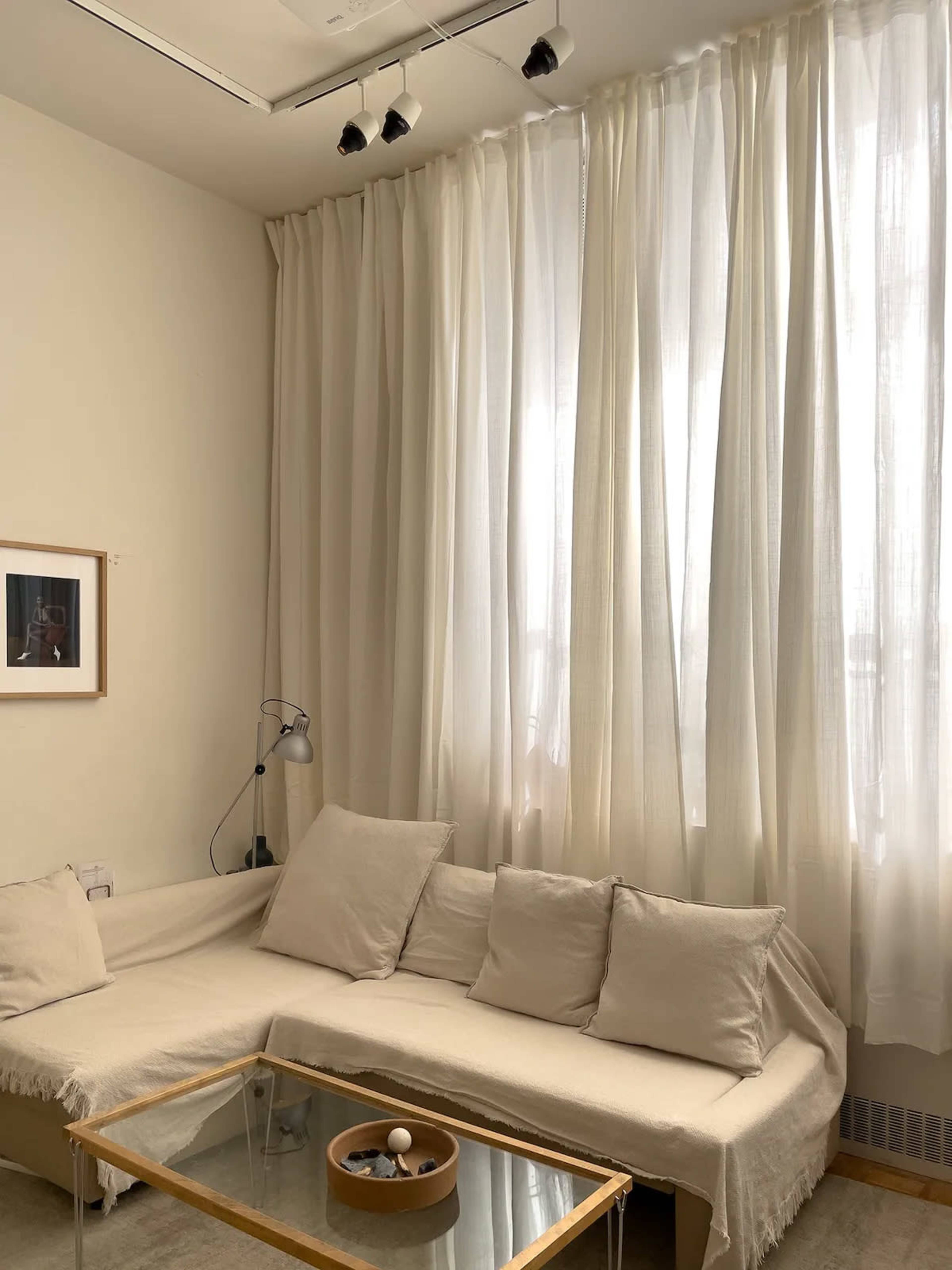 The image shows a cozy living room corner with a light-colored sofa, a glass coffee table, and sheer white curtains covering a window.