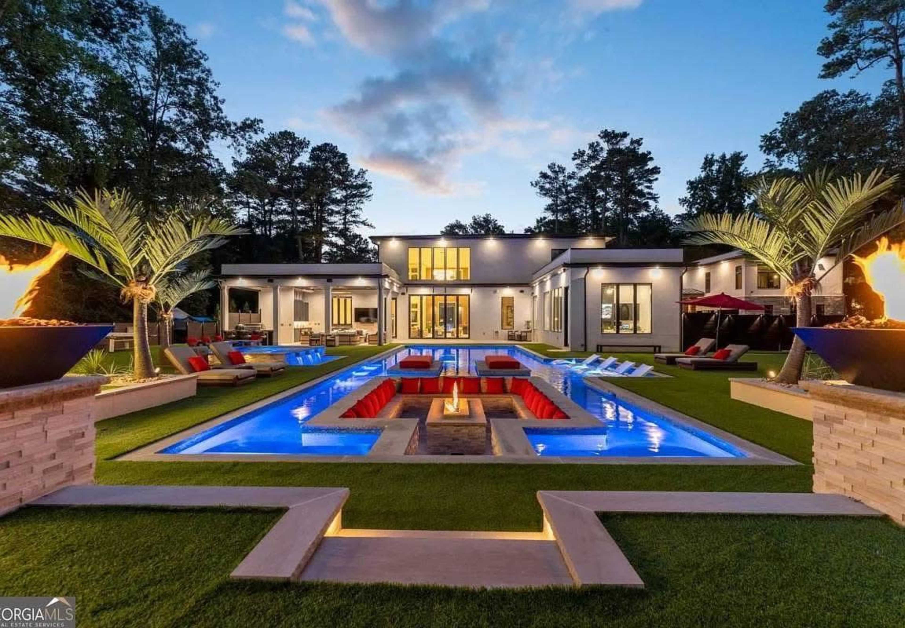 A modern luxury home with an illuminated swimming pool surrounded by palm trees and outdoor seating in a landscaped yard.