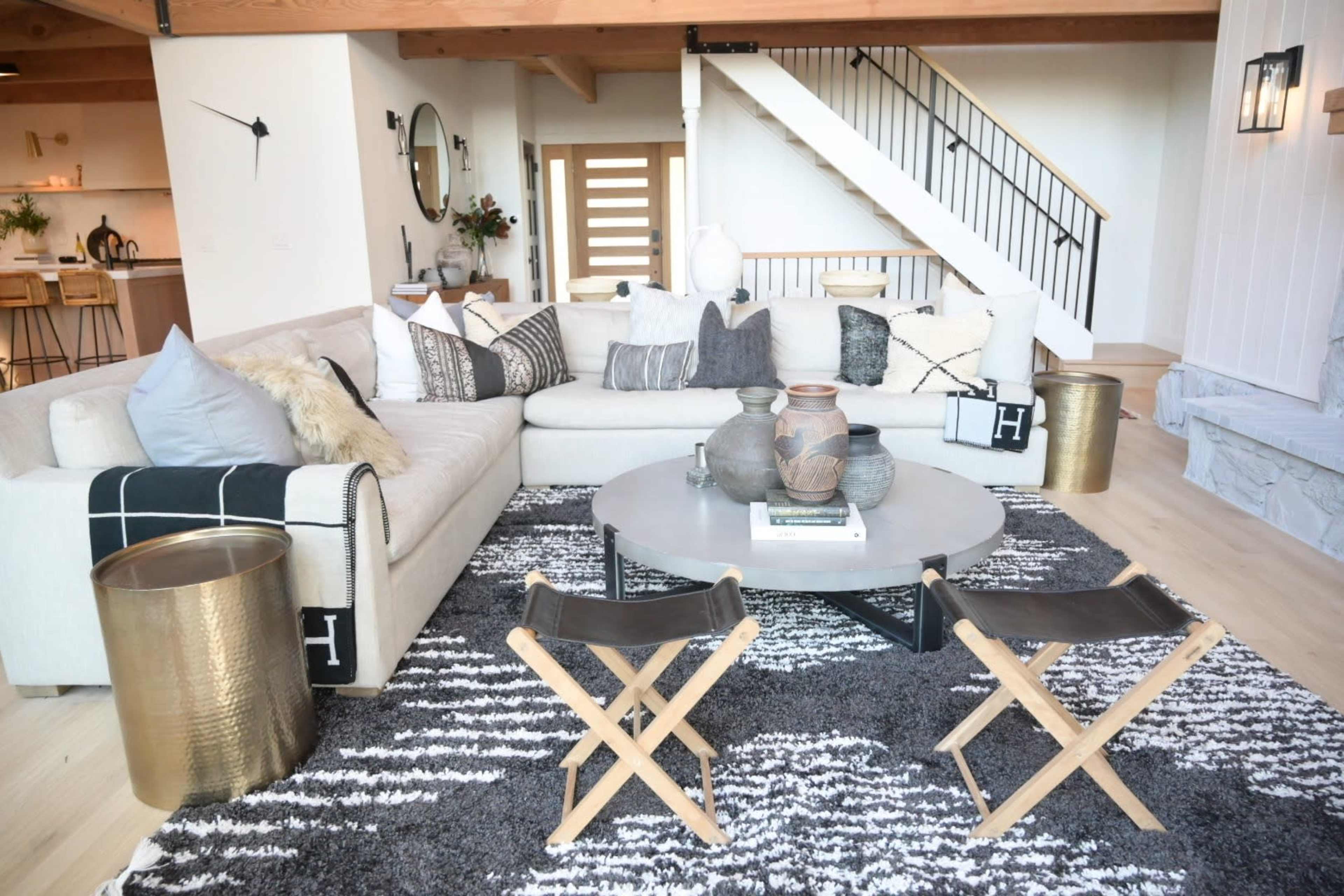 A modern living room featuring a large sectional sofa, a round coffee table with decorative items, and two folding chairs on a textured area rug under warm wood beams.