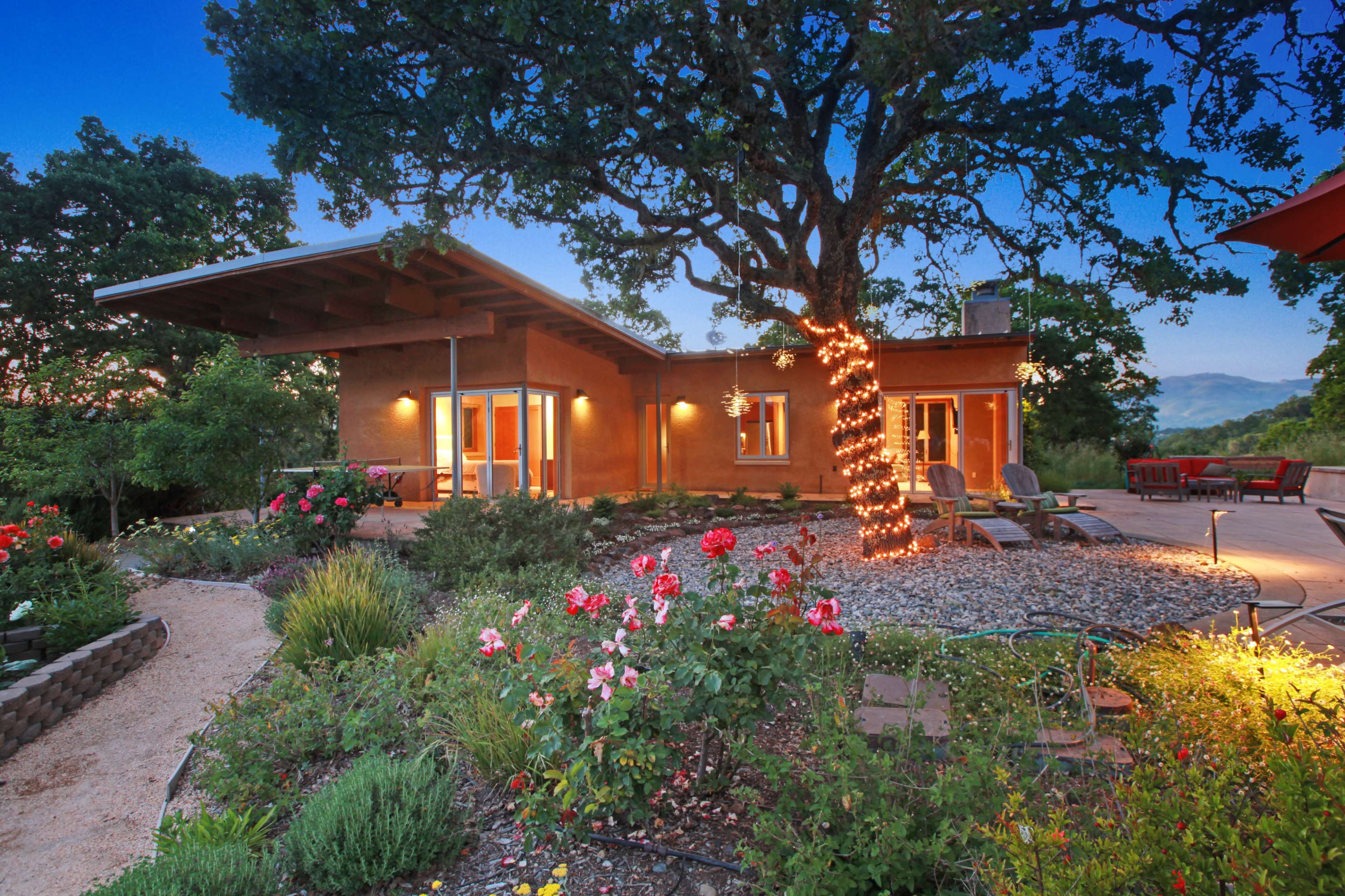 A modern adobe-style house surrounded by lush gardens and a tree wrapped in lights at twilight.
