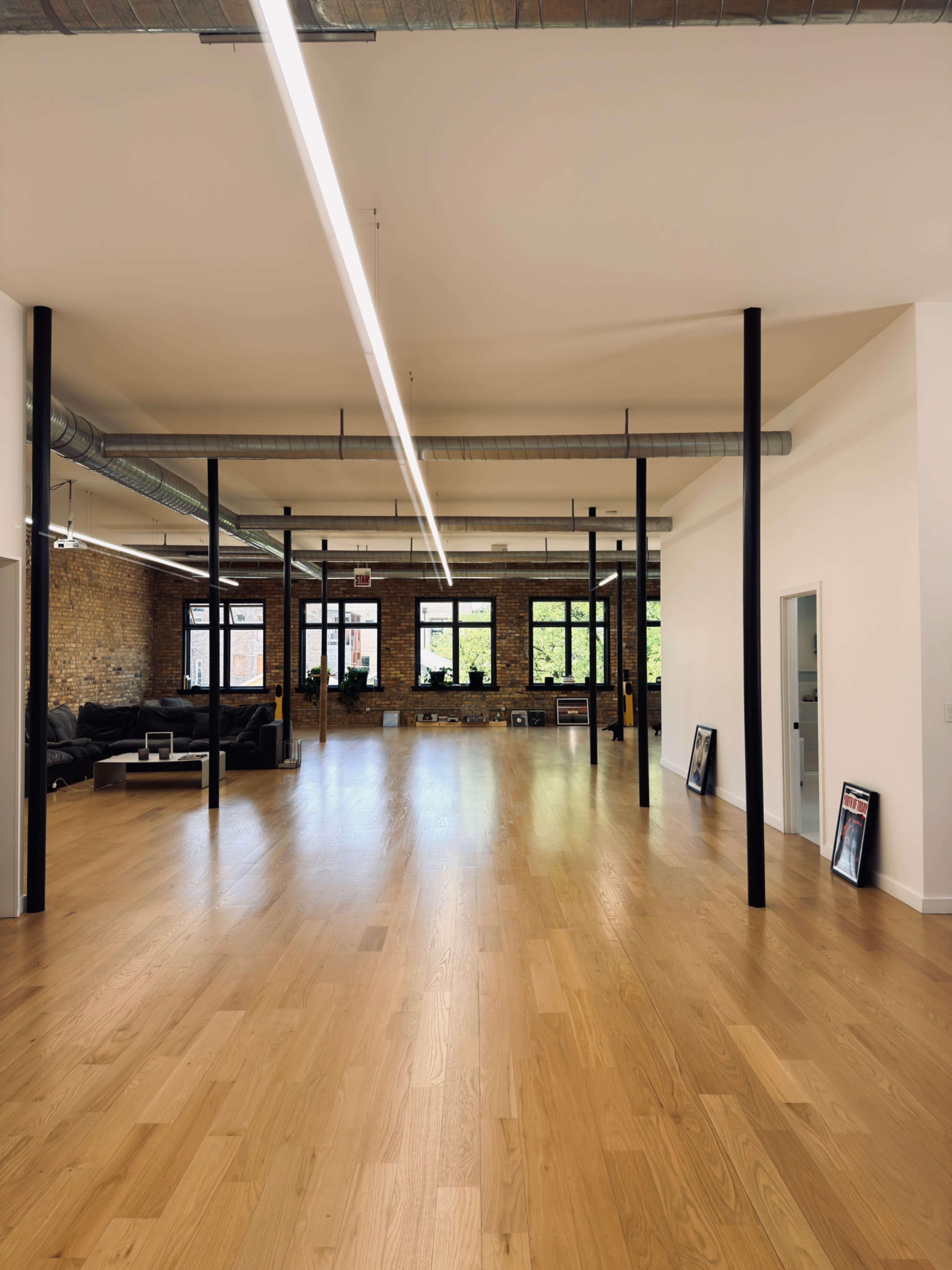The image depicts a spacious and modern interior of an office or studio with wooden flooring, large windows, and exposed brick walls.