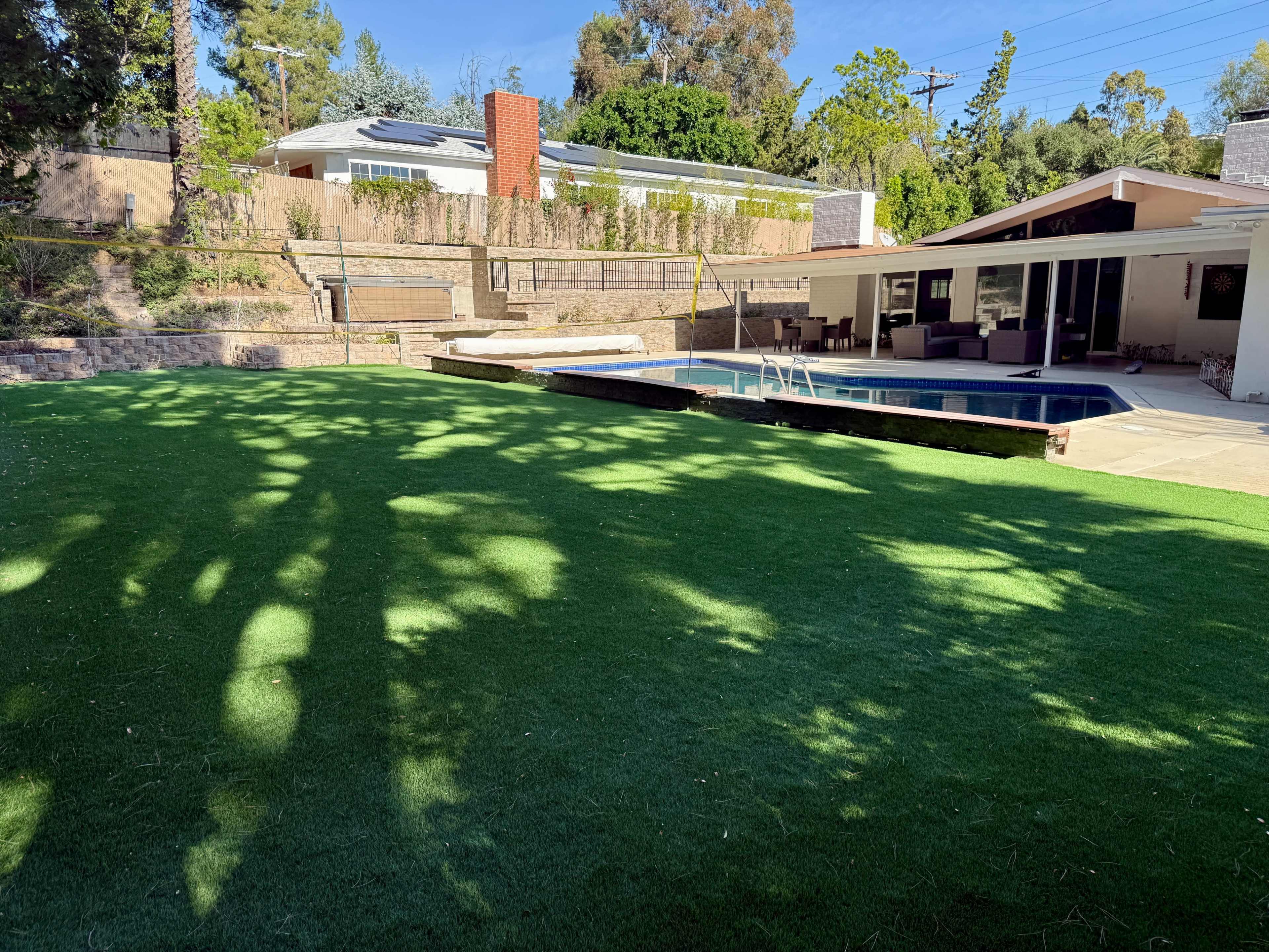 The image shows a yard with artificial turf, a swimming pool, and a modern house in the background surrounded by trees.