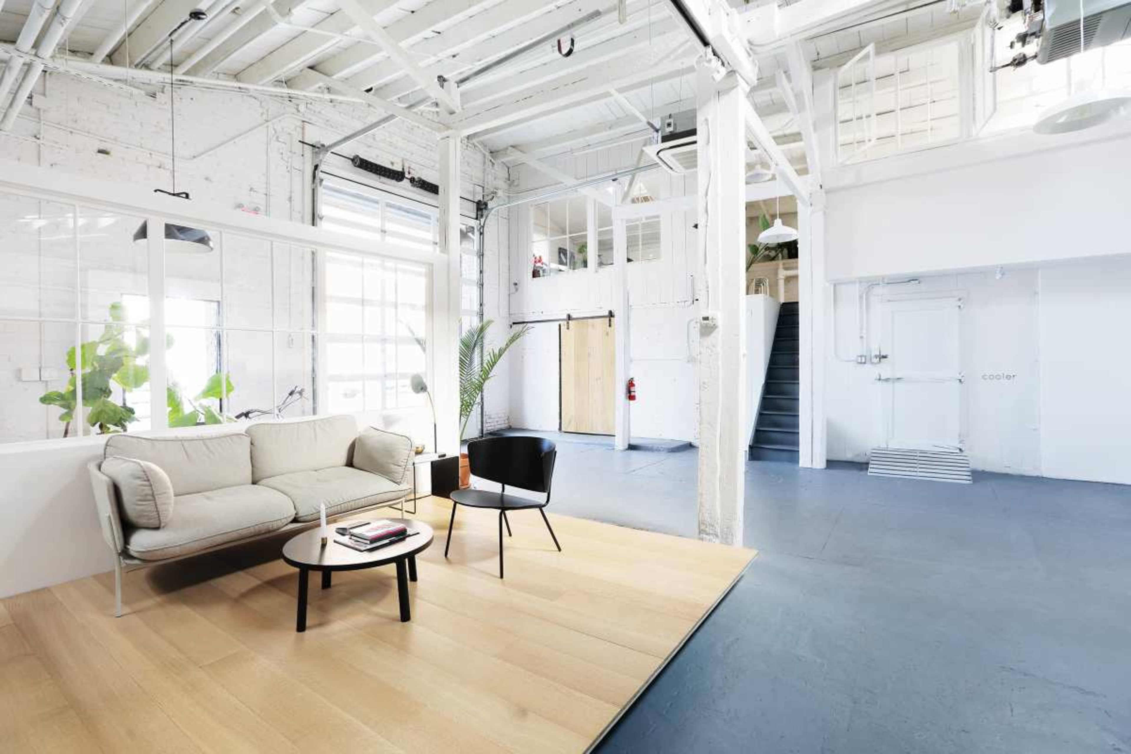 The image shows a spacious, modern interior with a sofa and a chair on a wooden platform, large windows, and a stairwell leading to an upper level.