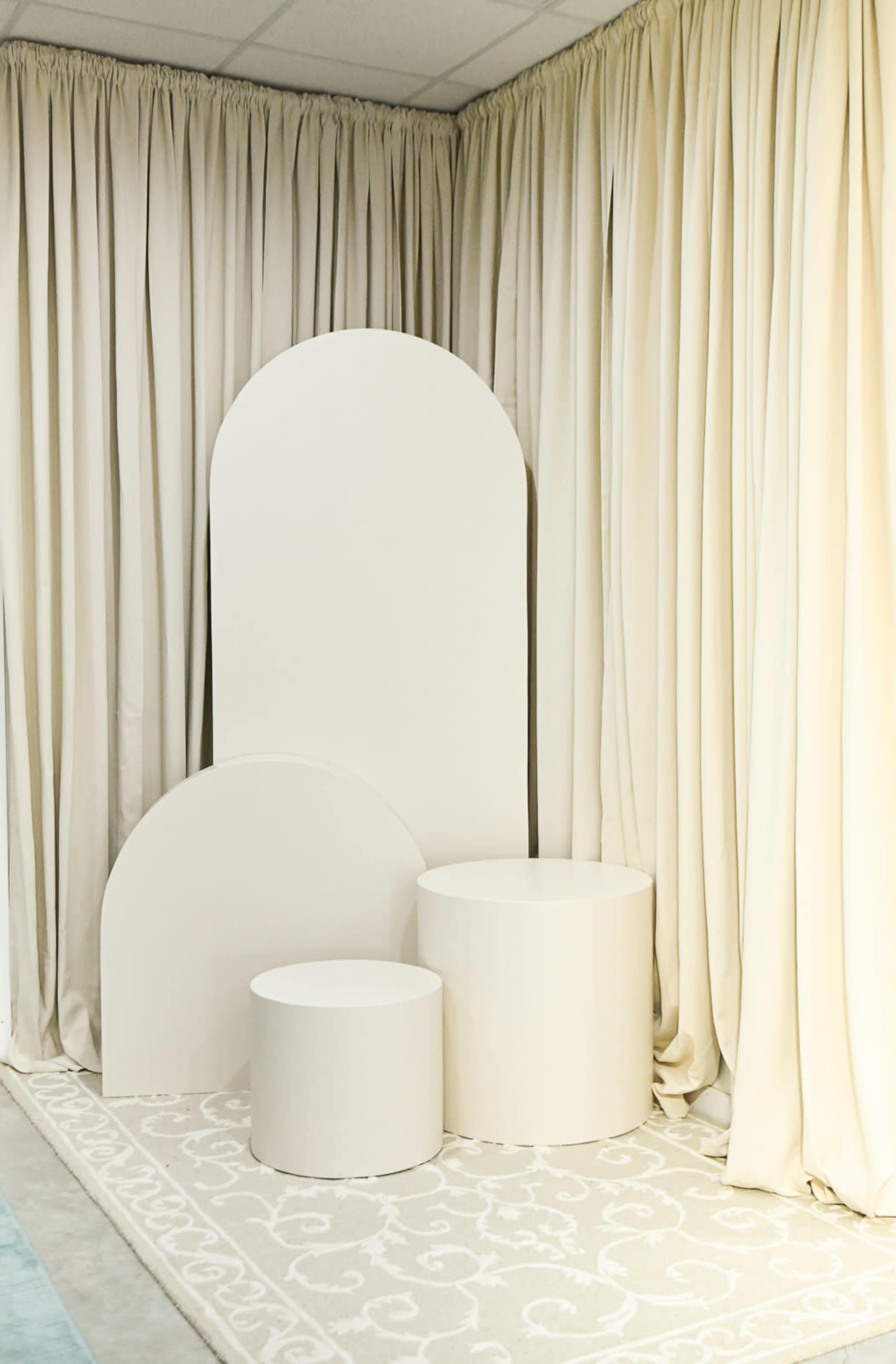 A minimalistic display featuring three curved white podiums against a backdrop of beige curtains.