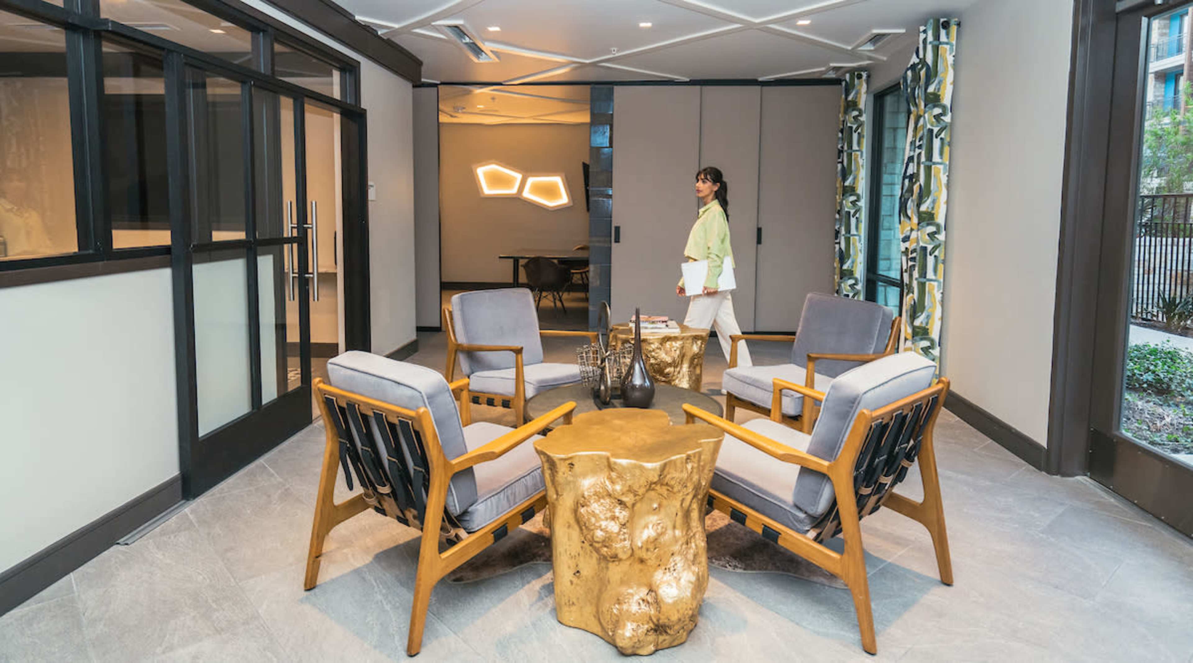 A woman walks through a modern lobby area featuring four wooden chairs, a gold accent table, and contemporary lighting.