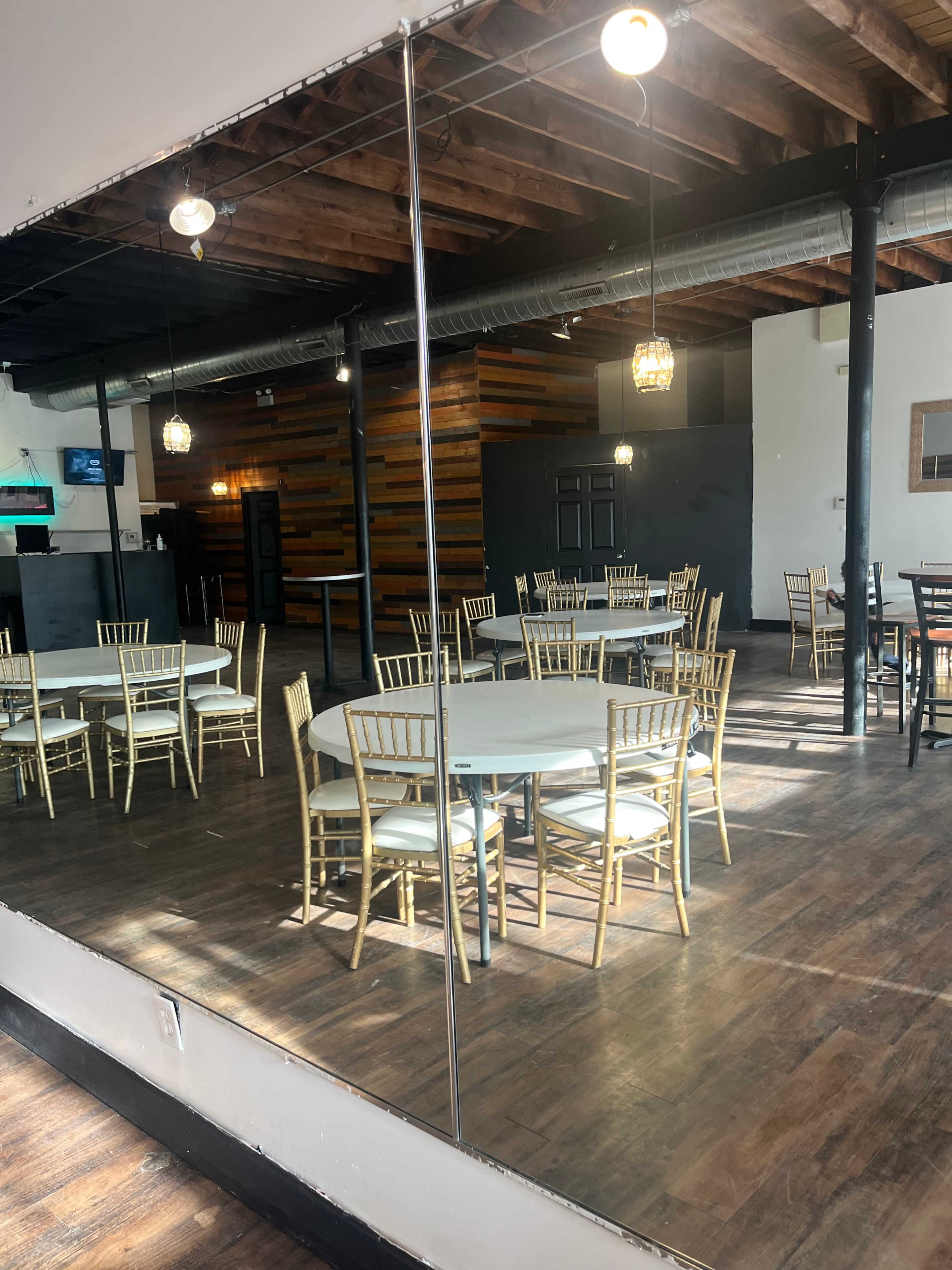 The image shows a spacious room with wooden floors, featuring several round tables surrounded by gold chairs, all reflected in a large mirror on one wall.