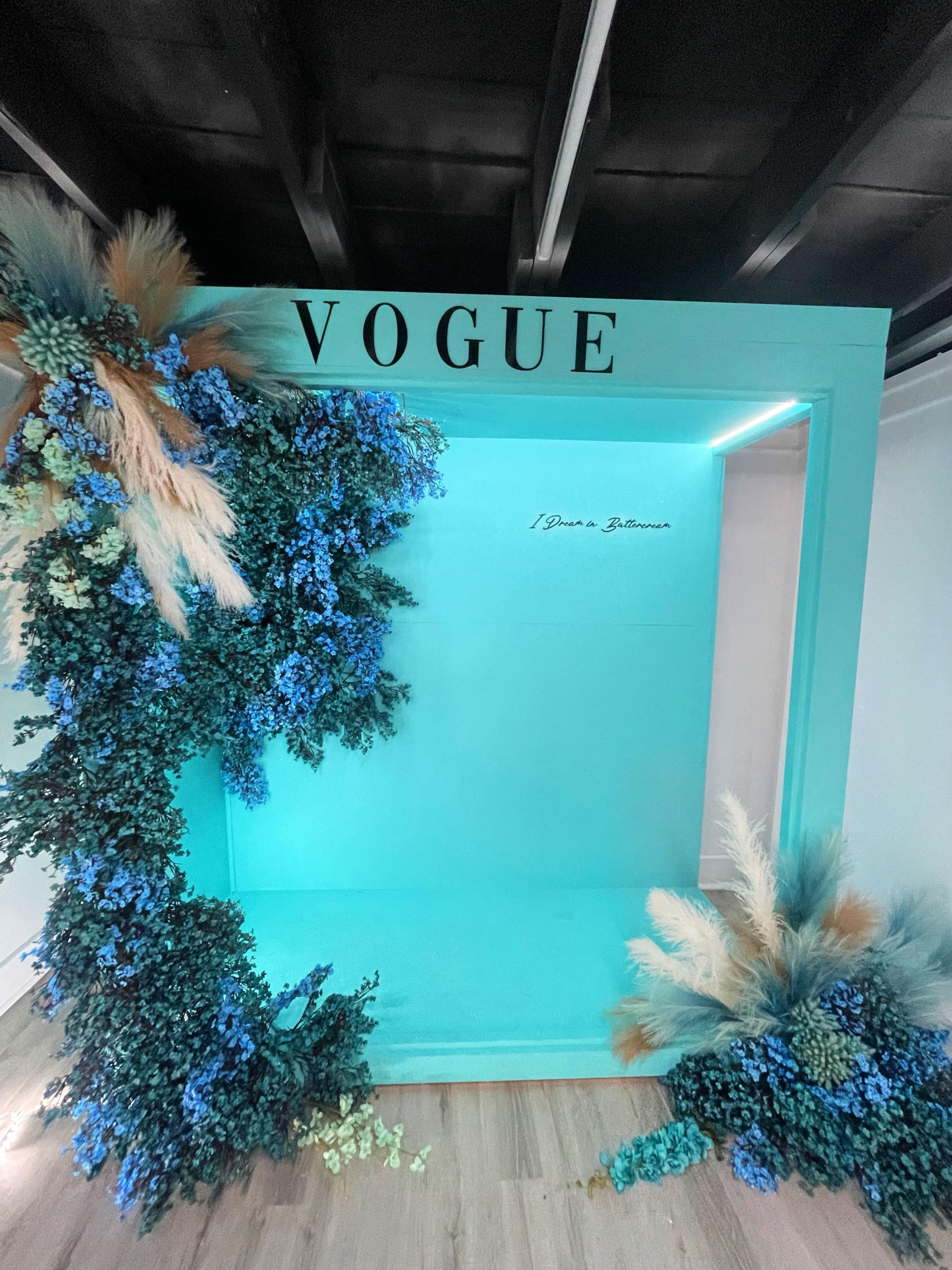 A vibrant display featuring a "VOGUE" sign at the top, surrounded by decorative blue and green foliage on one side.