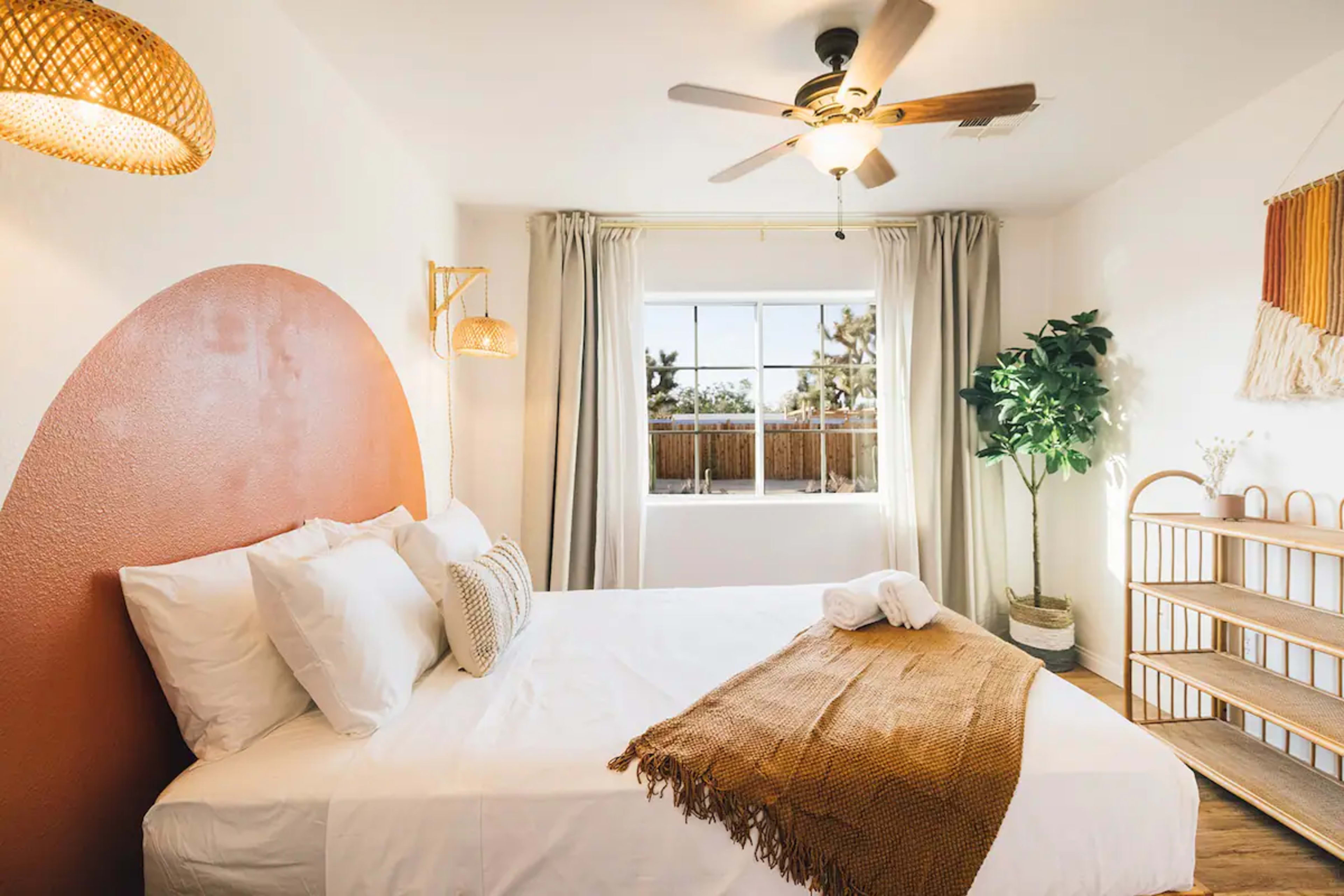 A bright bedroom featuring a large bed, a window with sheer curtains, a decorative plant, and a woven shelf.
