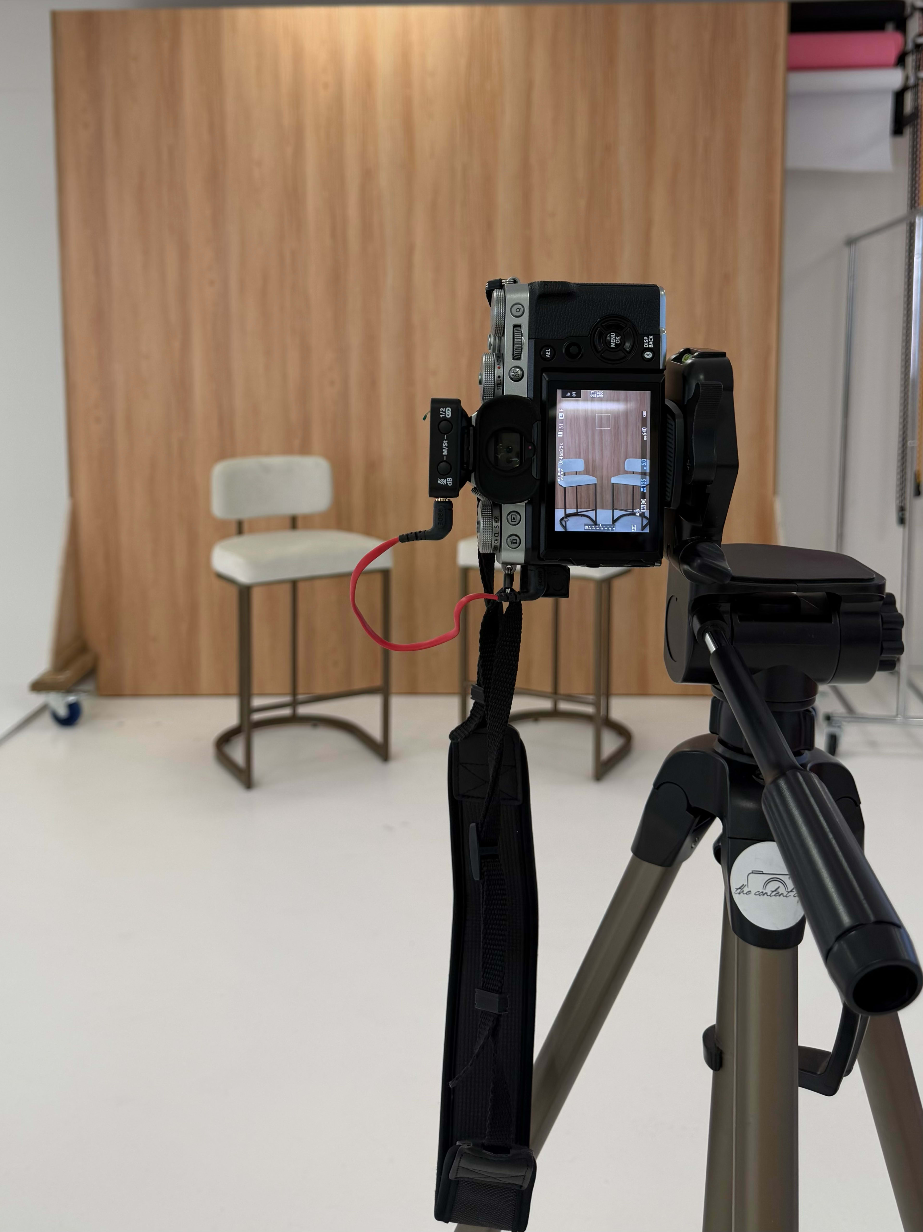 A camera on a tripod is positioned in front of a wooden backdrop, focused on a minimalist chair and a stool.