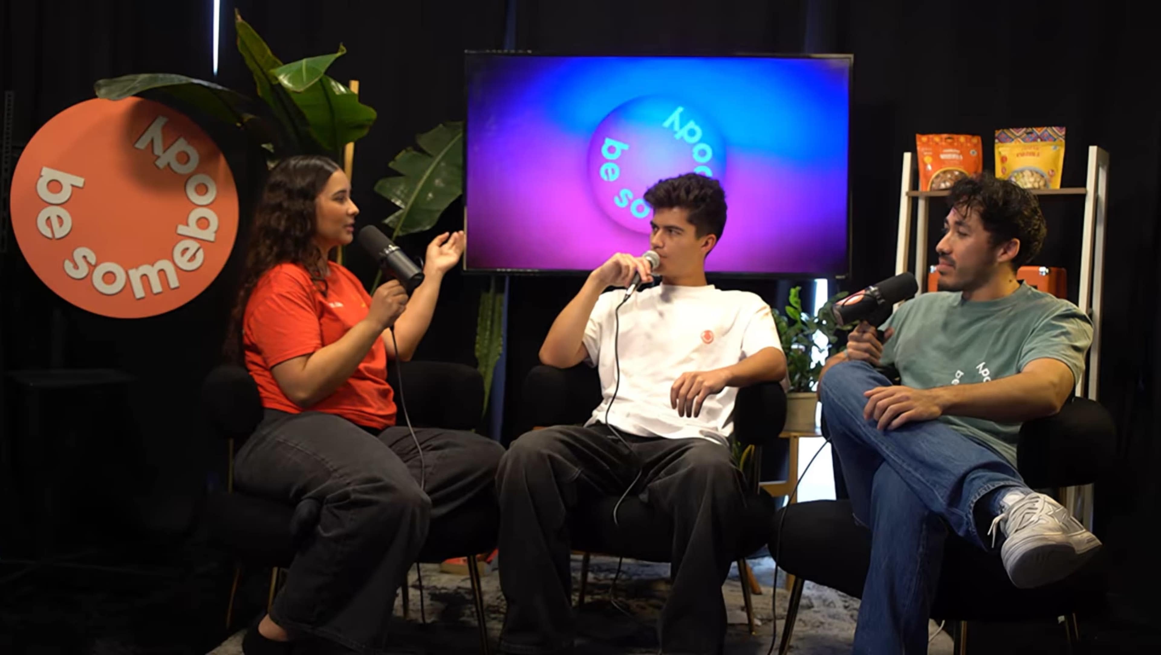 Three individuals are seated on black chairs in a studio setting, engaging in conversation while holding microphones and surrounded by plants and colorful decorations.