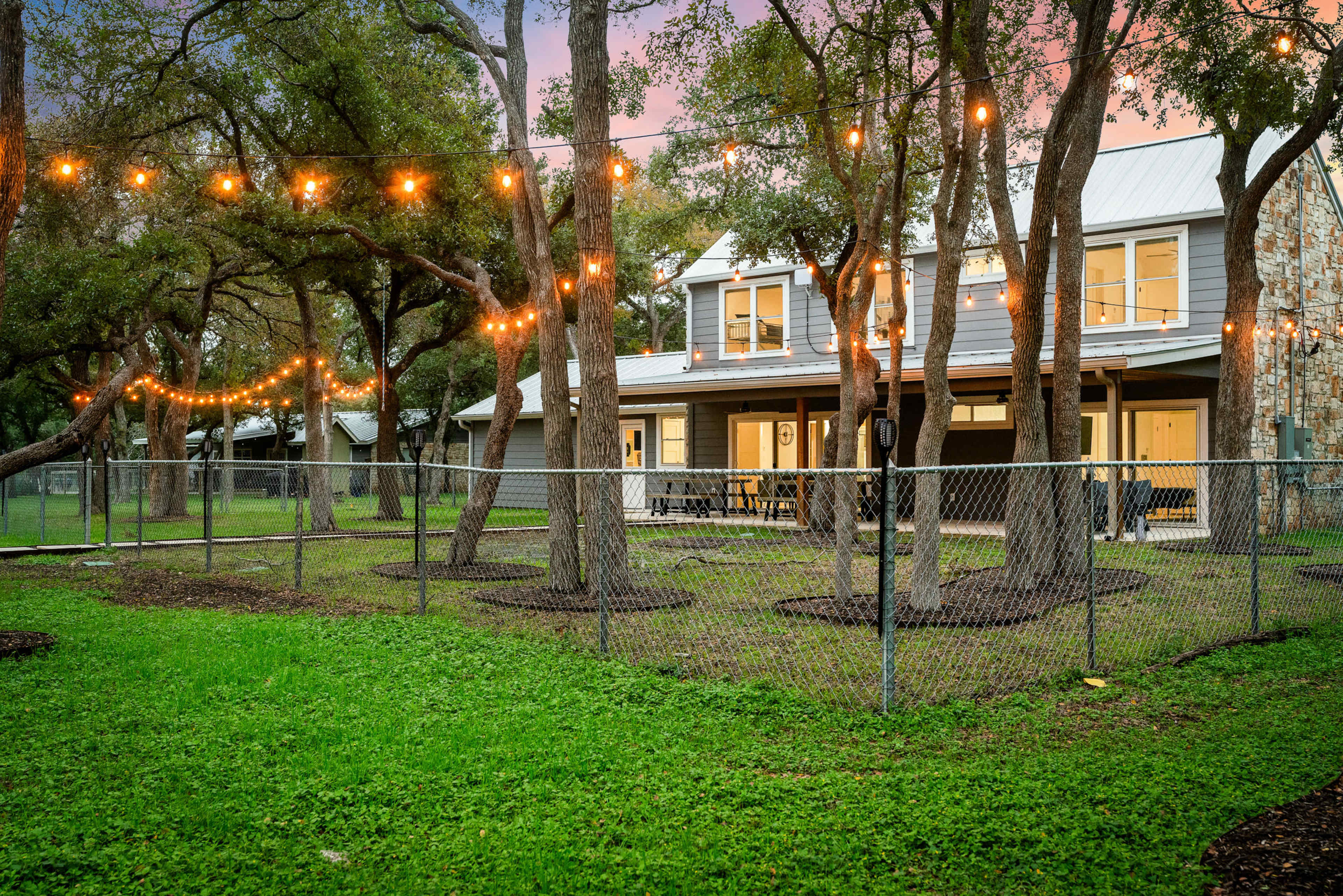 A modern house with a large yard and string lights is enclosed by a chain-link fence.