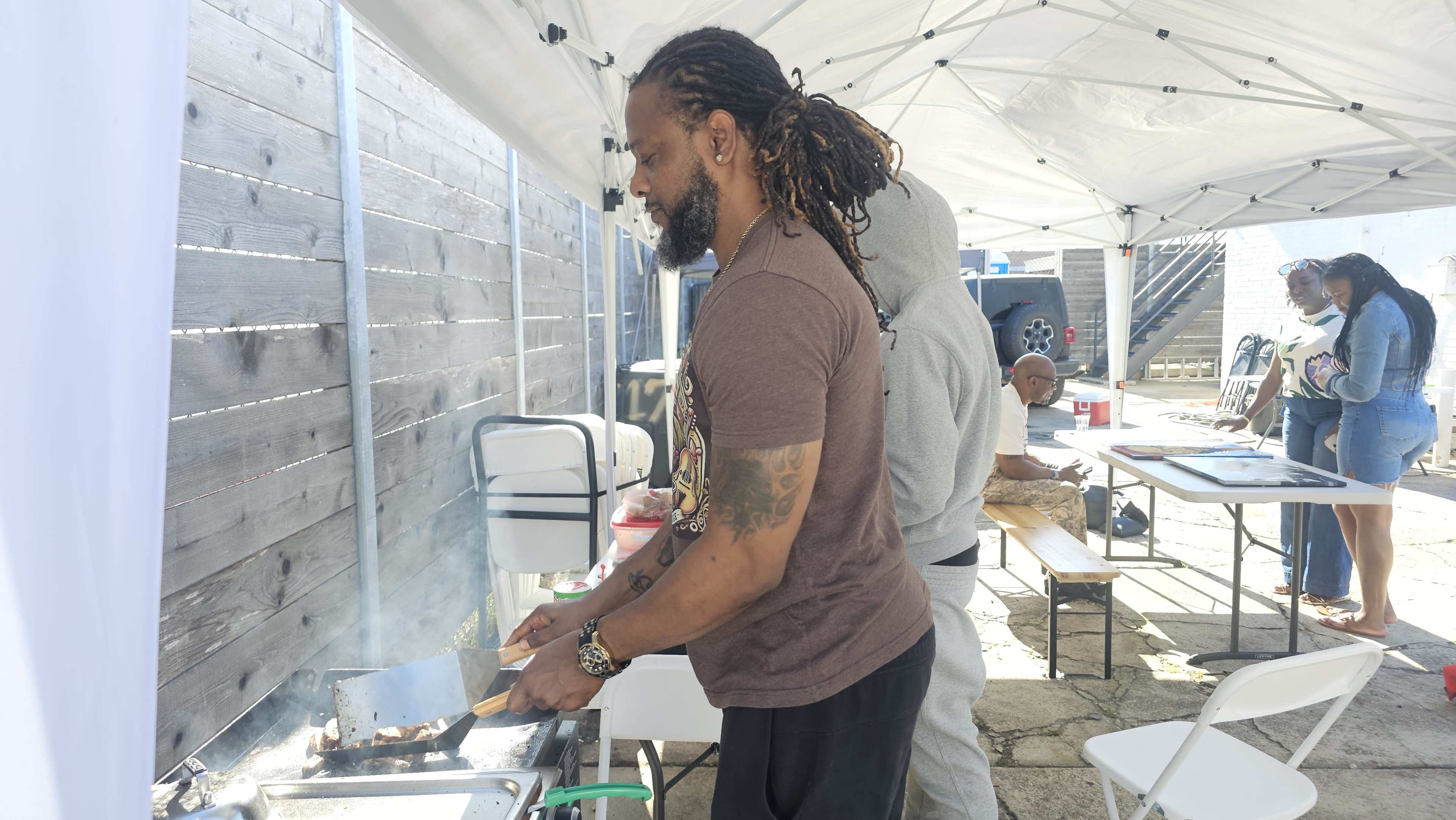 A man grills food outdoors while others engage in various activities under a white canopy.