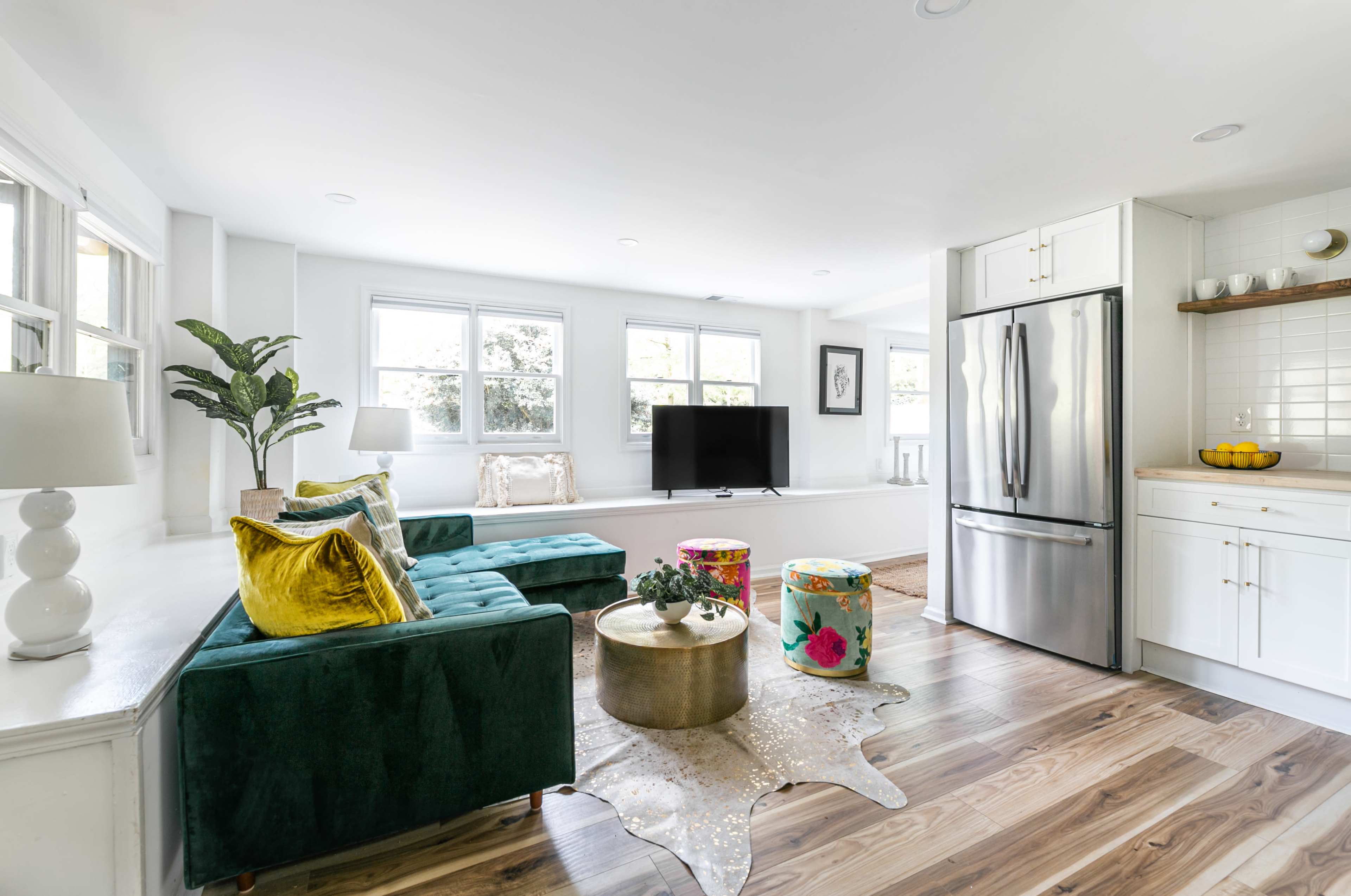 A modern living space features a green sectional sofa, a gold coffee table, colorful stools, and a kitchen area with stainless steel appliances.