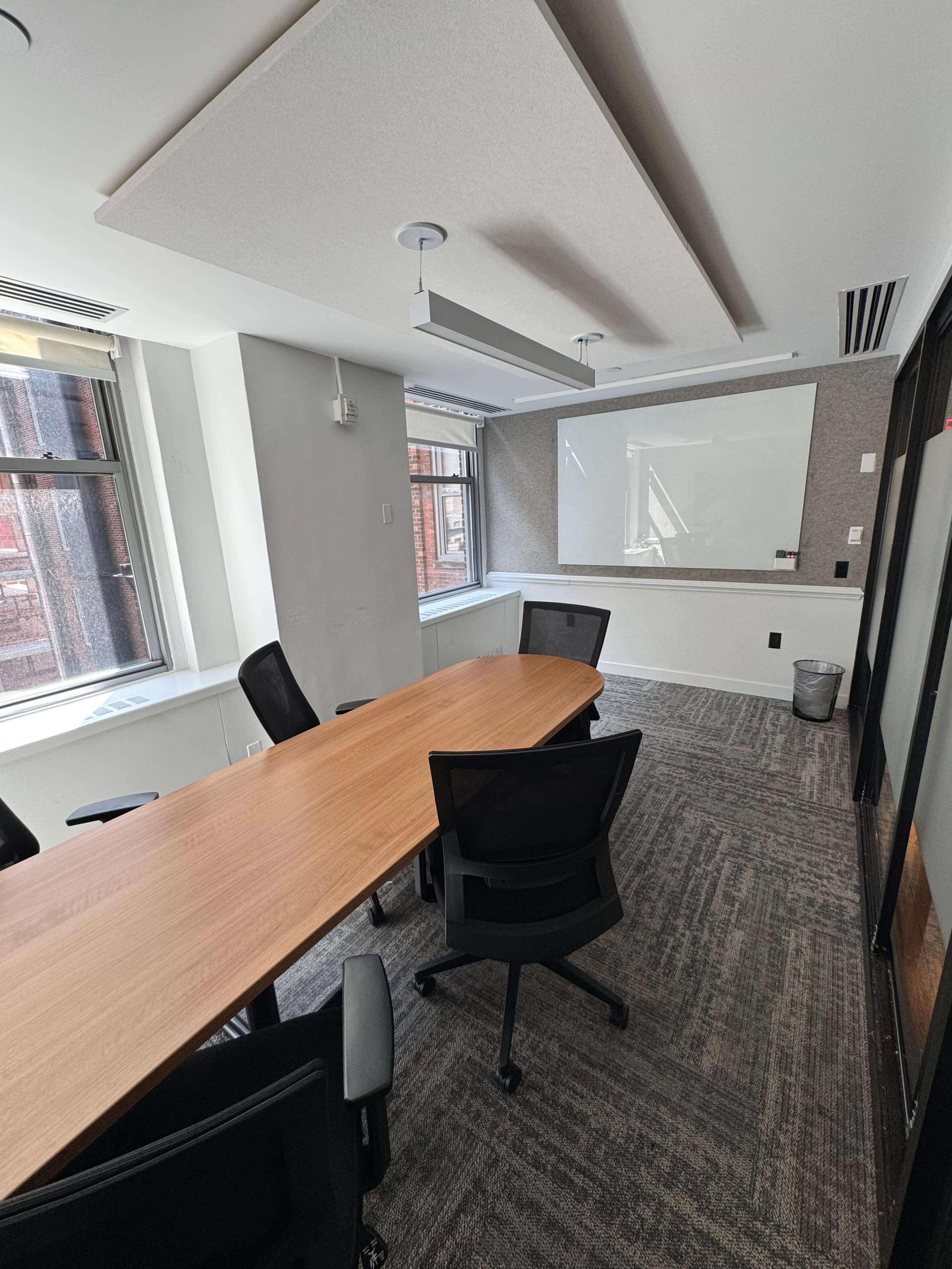 A modern conference room features a long wooden table surrounded by black chairs, with large windows and a whiteboard on the wall.
