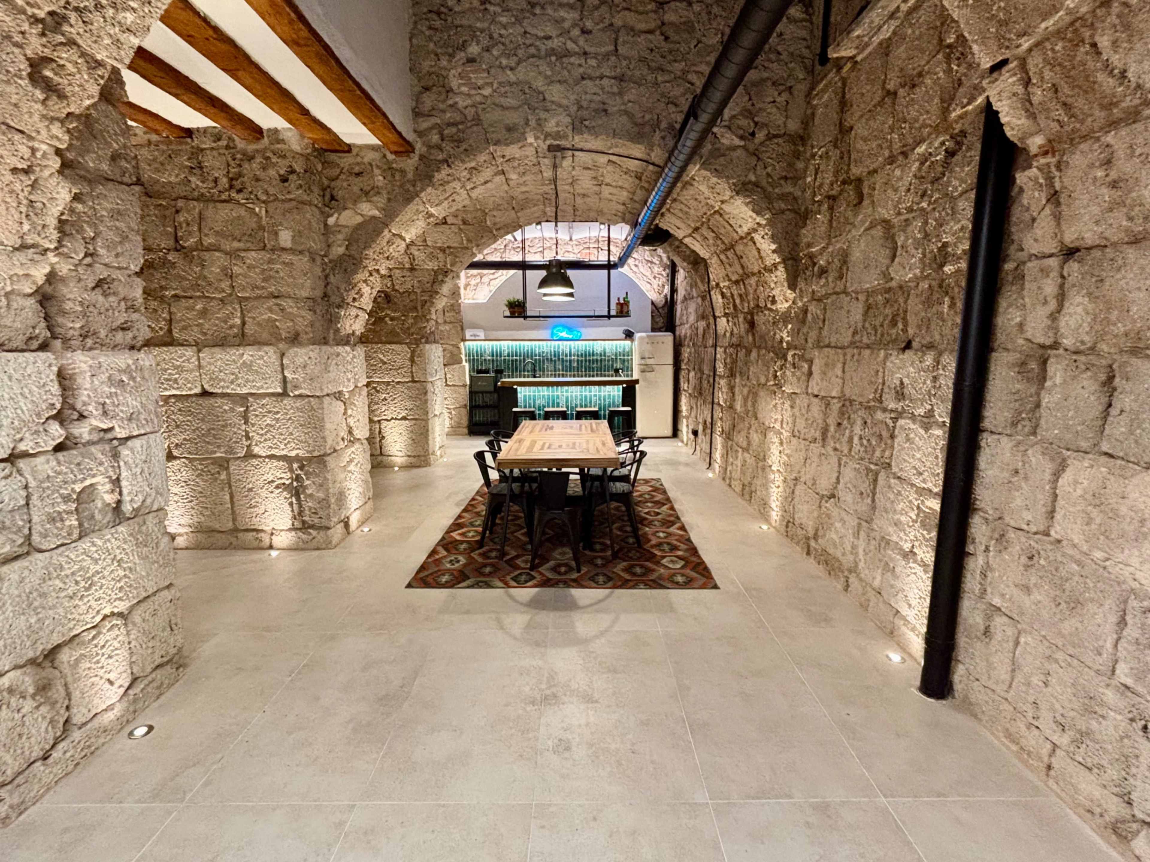 The image shows a spacious, stone-walled room with arched doorways, featuring a dining table surrounded by chairs and a kitchen area in the background.