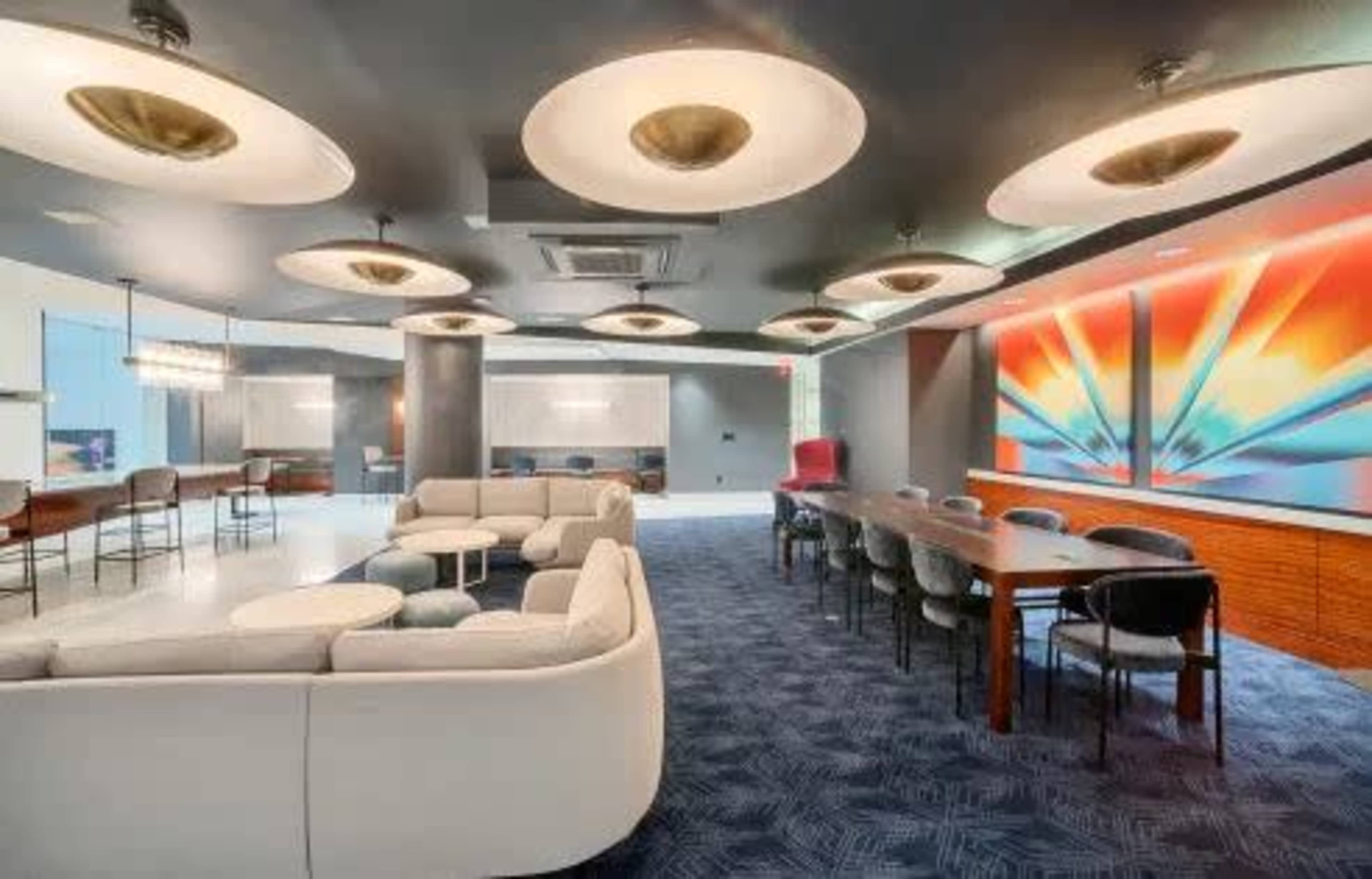 The image shows a modern lounge area featuring a seating arrangement with sofas and coffee tables, a long dining table, and artistic wall décor illuminated by circular ceiling lights.