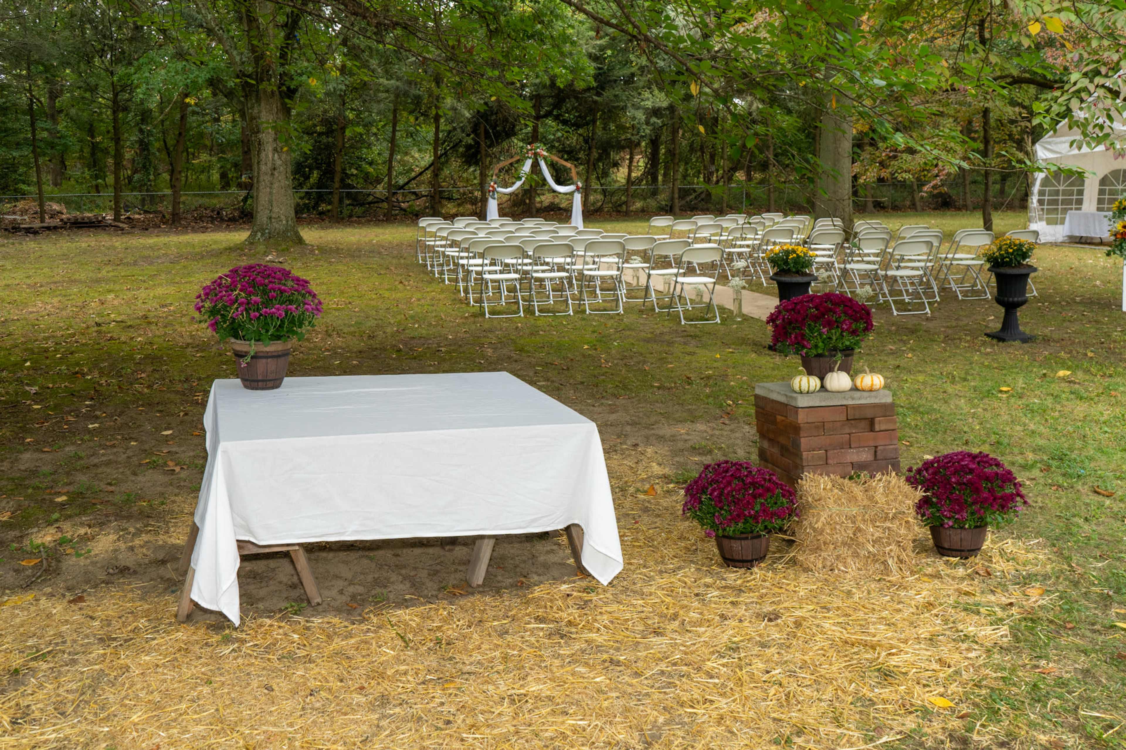 A table draped with a white cloth is set beside potted flowers and hay bales, with rows of folding chairs arranged for a gathering in a wooded area.