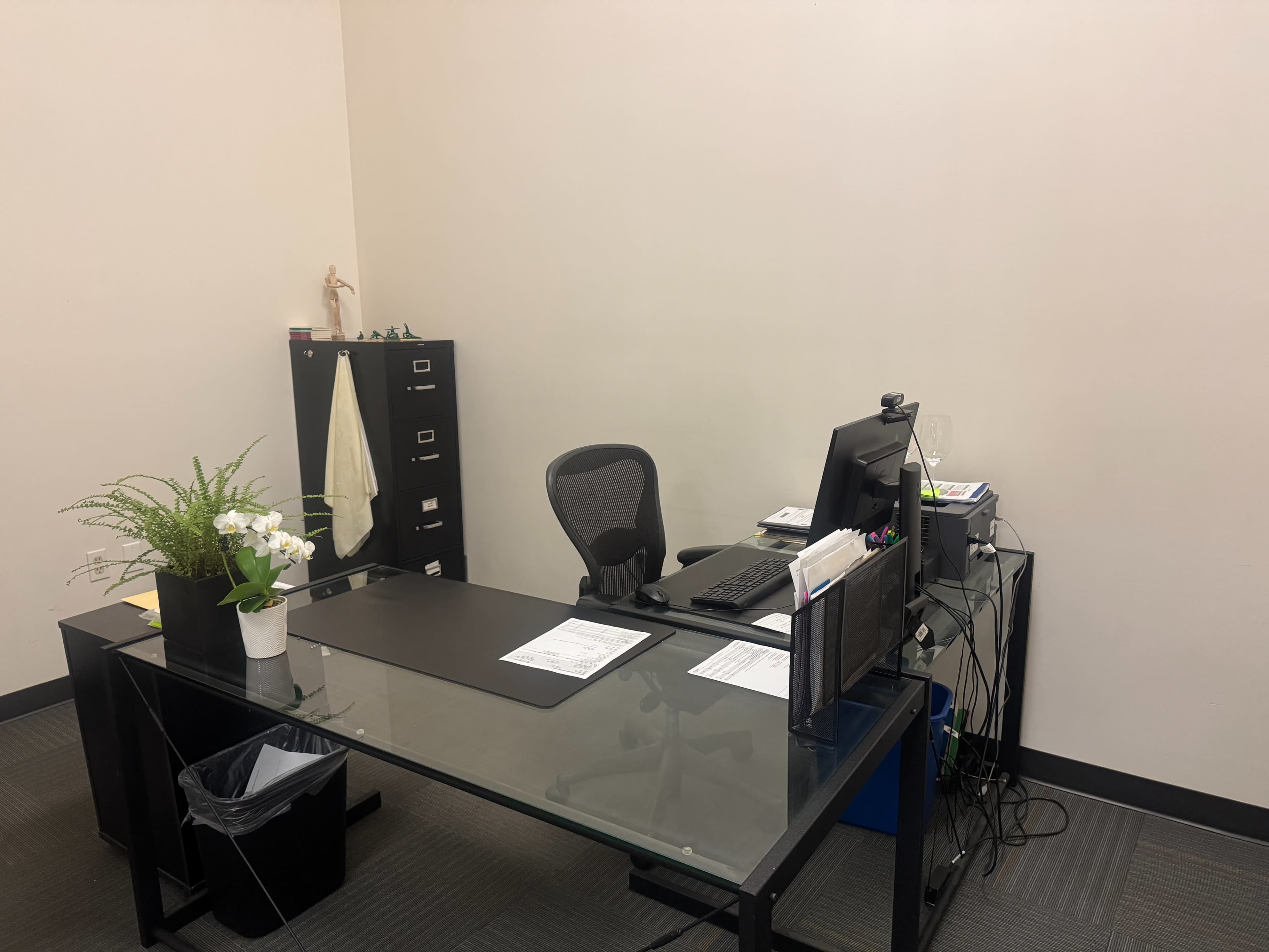 The image shows a minimalist office with a glass-top desk, a black office chair, a computer, and a filing cabinet against a plain wall.