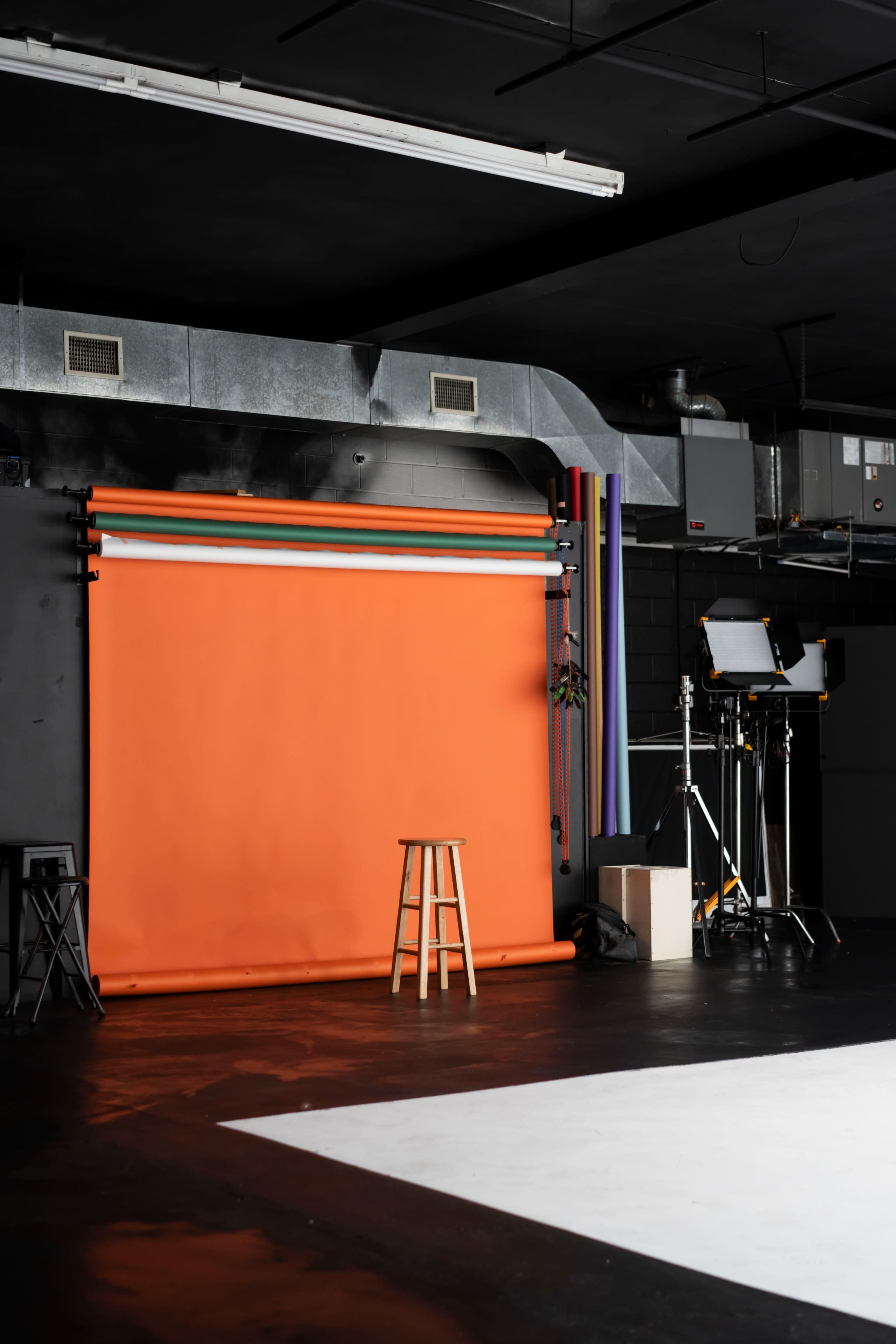 The image shows a photography studio with an orange backdrop, a wooden stool, and professional lighting equipment.