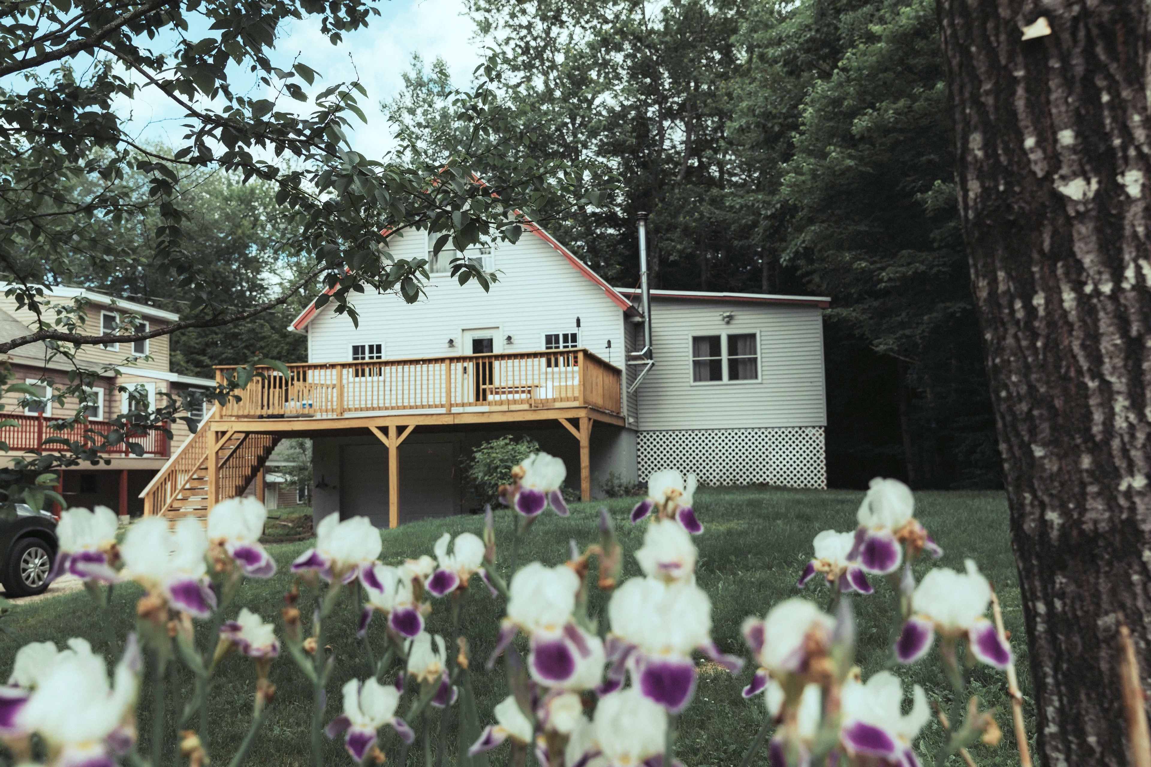 A two-story house with a wooden deck is surrounded by green grass and blooming purple and white flowers.